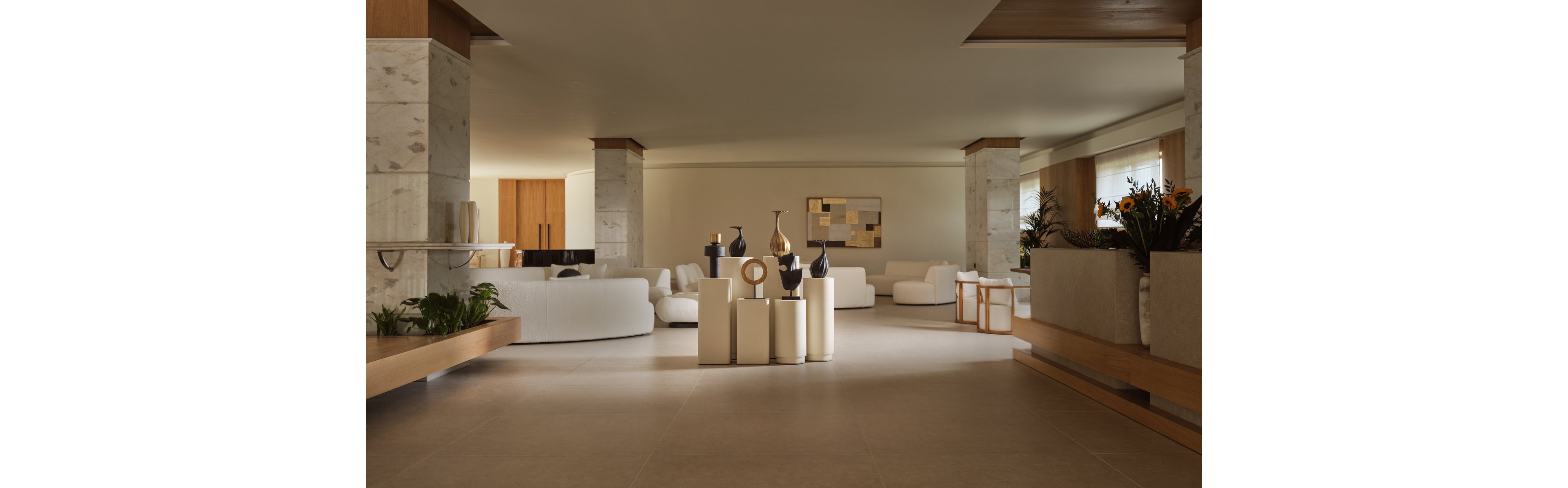 A large contemporary interior features a spacious lobby or lounge area with minimalist white sofas and armchairs. Tall marble-clad columns divide the open-plan space, while sculptural vases and candles are arranged on cylindrical pedestals in the center. Warm neutral tones dominate the room, complemented by wooden accents and soft, indirect lighting. Potted plants and a modern abstract wall artwork add texture and visual interest to the serene setting.