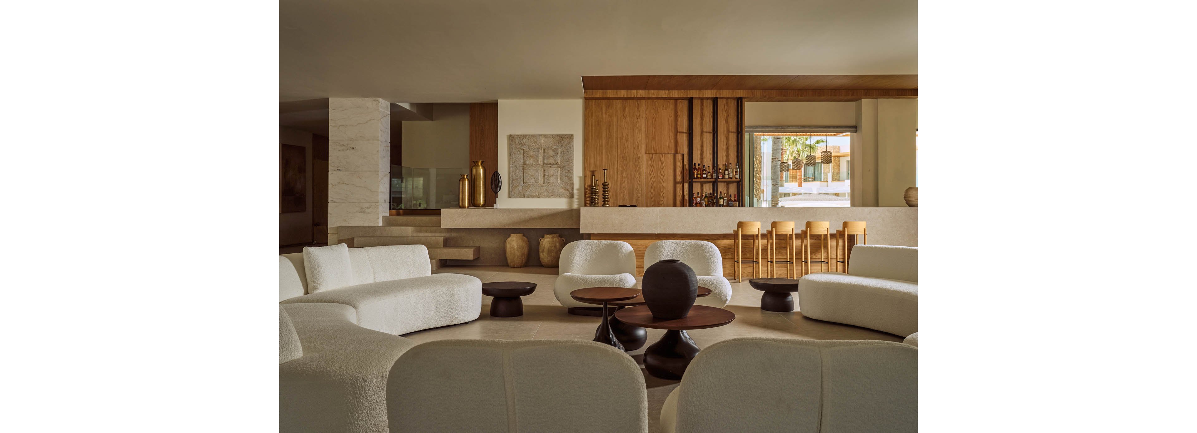 A contemporary lounge features curved white sofas arranged around low dark coffee tables. In the background, a wooden bar with high stools and decorative vases creates a warm focal point. Neutral tones of beige, cream, and wood dominate the space, giving it a minimalist yet inviting feel. Large windows near the bar bring in natural light and a glimpse of the outdoors.