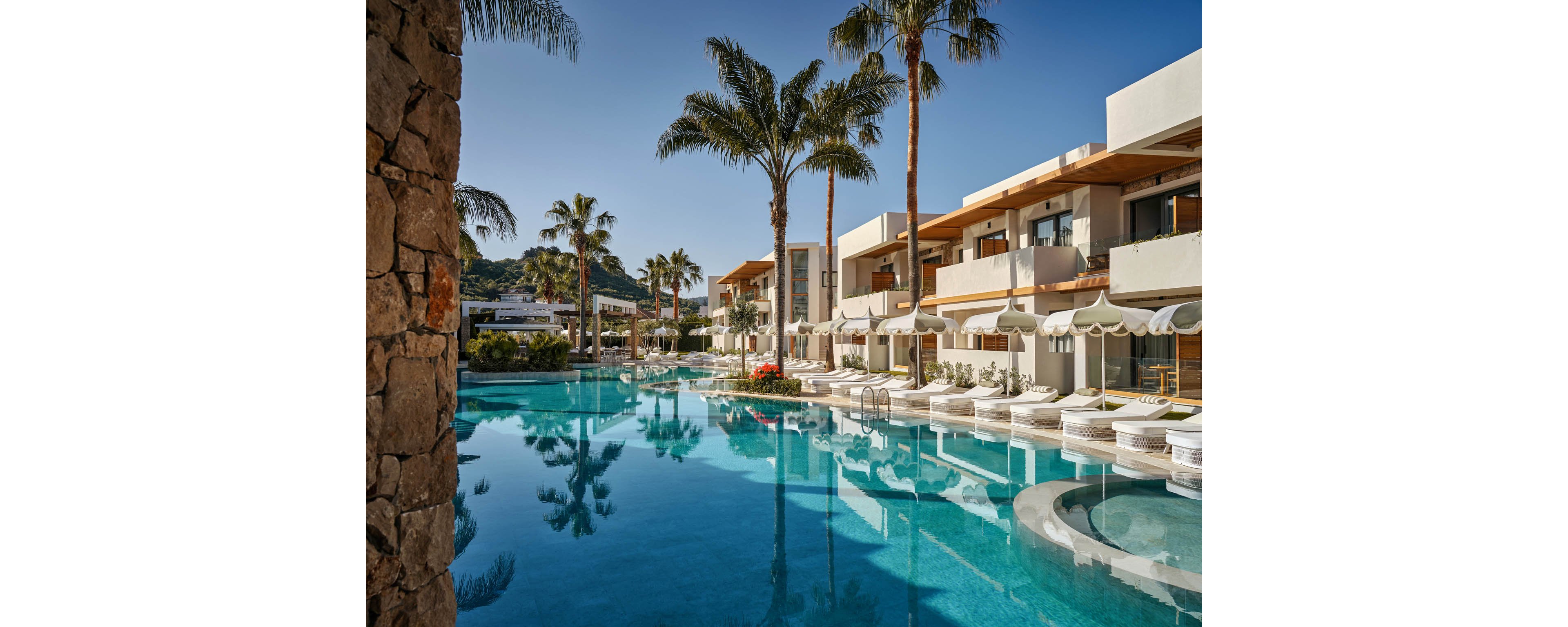 A luxurious resort pool stretches alongside modern white villas with wooden accents. Tall palm trees and surrounding mountains are reflected in the calm turquoise water. White sun loungers and umbrellas line the poolside, creating an inviting and serene atmosphere. The scene suggests a peaceful vacation destination in a warm, coastal climate.