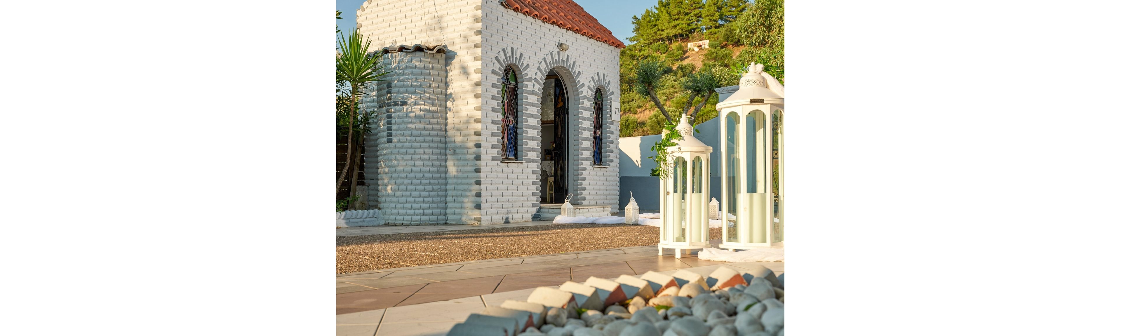 A small white brick chapel with arched stained-glass windows stands in a sunlit outdoor courtyard. Large white lanterns with candles are arranged in the foreground, adding a decorative touch to the scene. The ground is covered with smooth stones and tiled pathways leading toward the chapel entrance. Green trees and shrubs surround the area, creating a peaceful Mediterranean atmosphere.