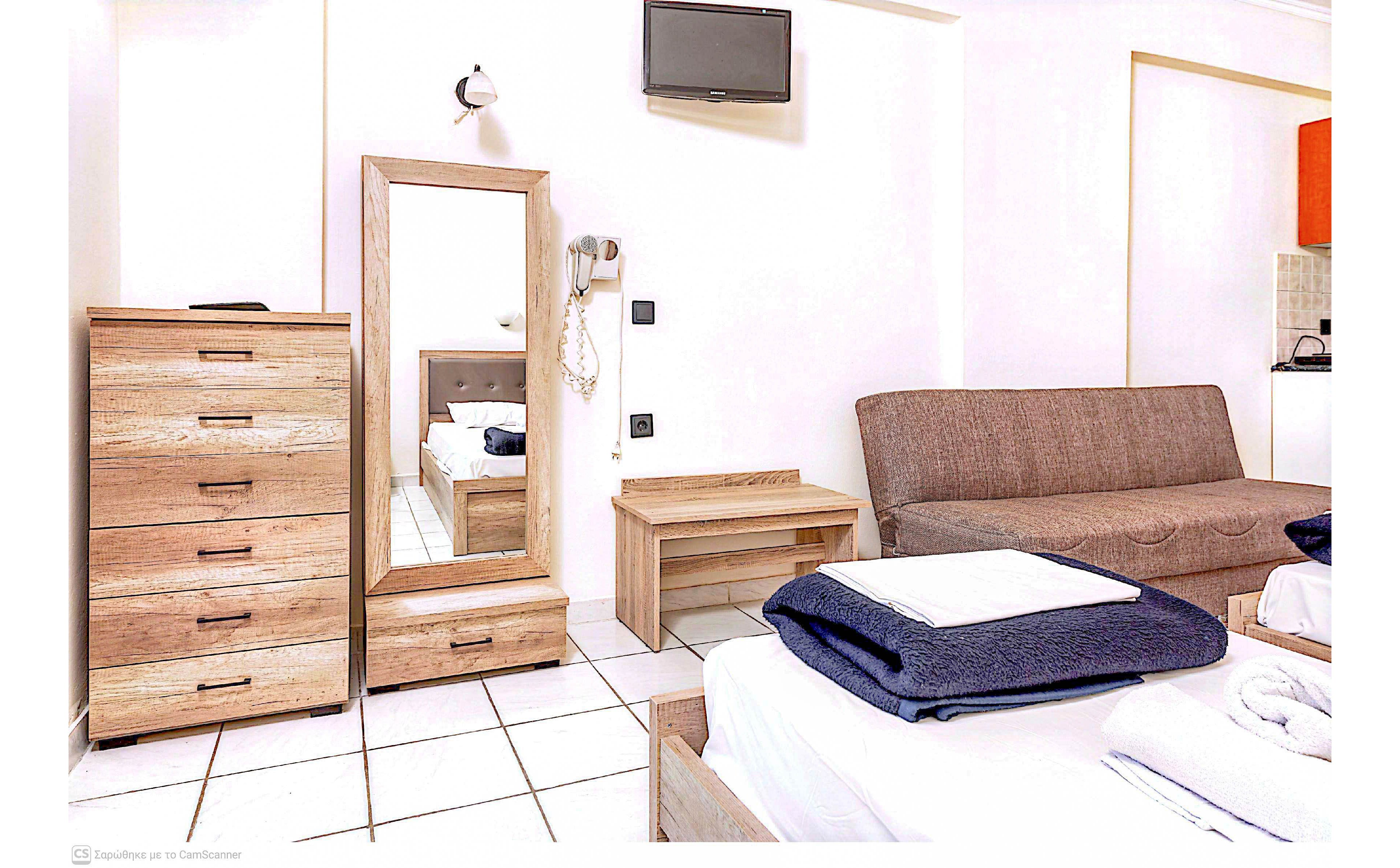 A clean, bright hotel room features a bed in the foreground with neatly folded white linens and a dark blanket. Along the wall, there is a wooden dresser, a tall mirror, and a small wooden table beside a brown sofa. A small wall-mounted television and light fixture are visible above the furniture, with white tiled flooring adding to the minimalist look. The overall setting feels simple, modern, and functional, suitable for short stays or vacation rentals.