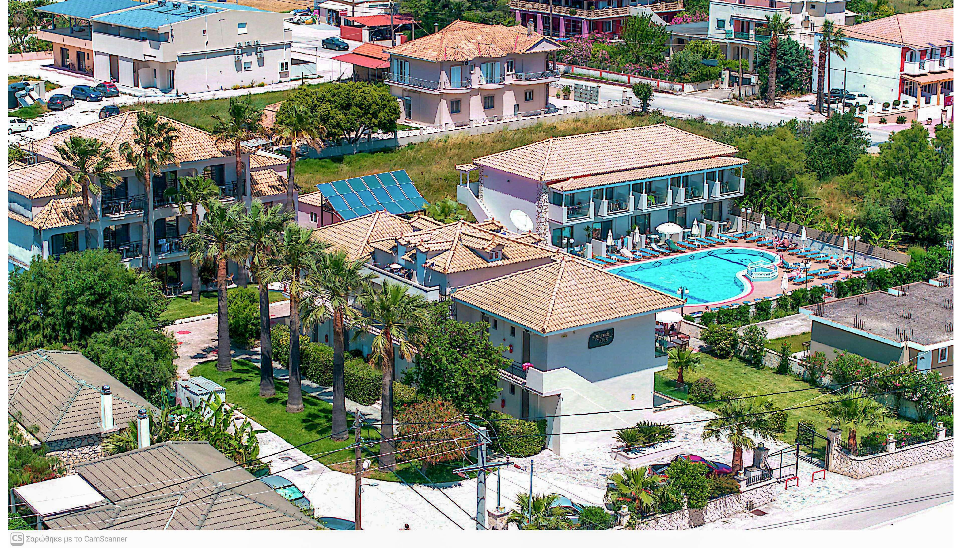 A seaside resort complex is captured from above, showing several low-rise buildings with tiled roofs. At the center is an outdoor swimming pool surrounded by lounge chairs and umbrellas. Palm trees and landscaped greenery line the property, with nearby roads and additional houses visible in the background. The bright daylight and clear weather create a vibrant, summery atmosphere.