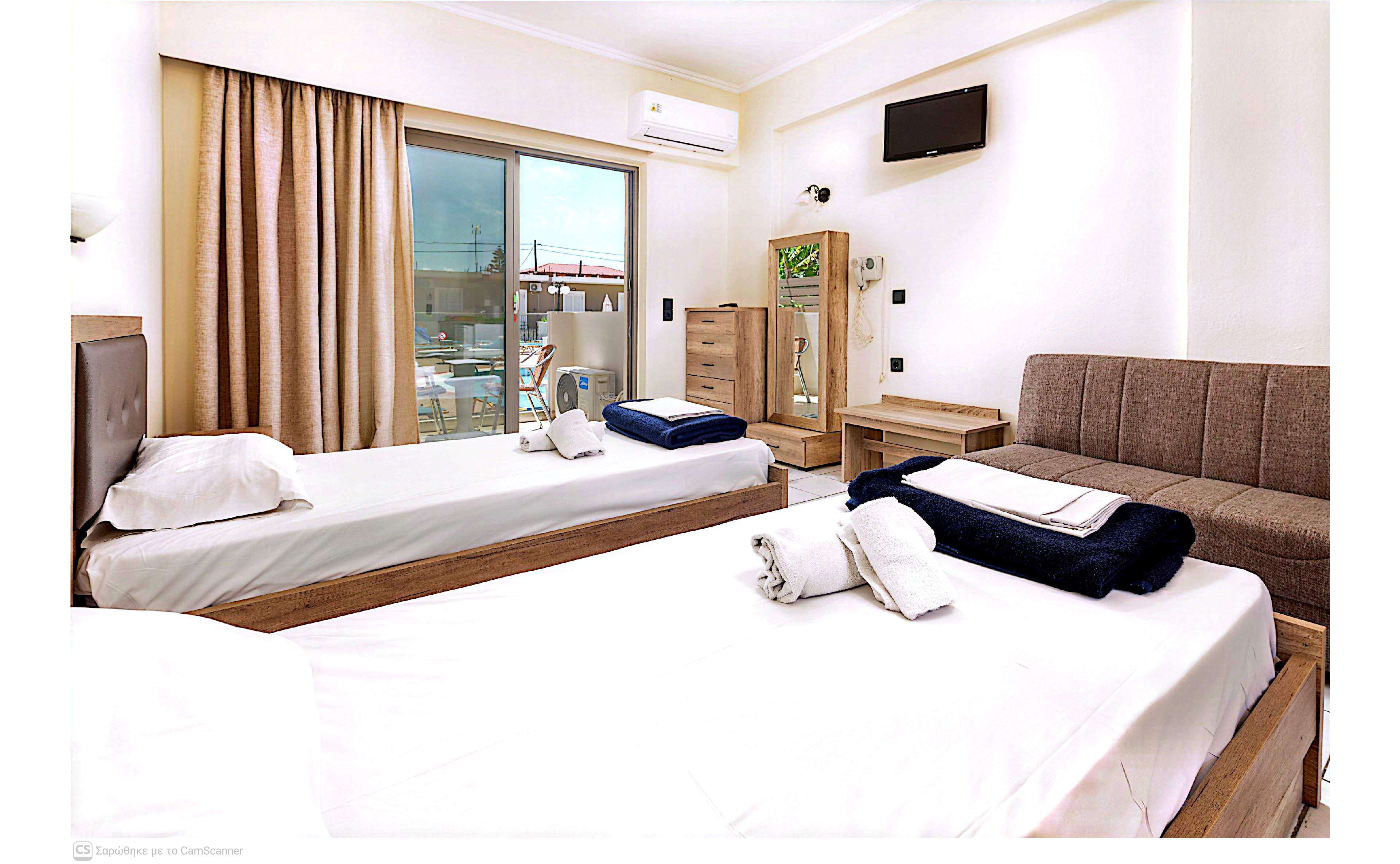 Interior view of a modern hotel room featuring two neatly made twin beds with folded towels and dark blue blankets. A sofa sits against the wall opposite a wall-mounted television, with a wooden dresser and mirror nearby. Large sliding glass doors lead to a balcony with outdoor seating and a cityscape visible beyond. Neutral walls and light wood furniture create a clean, contemporary atmosphere.