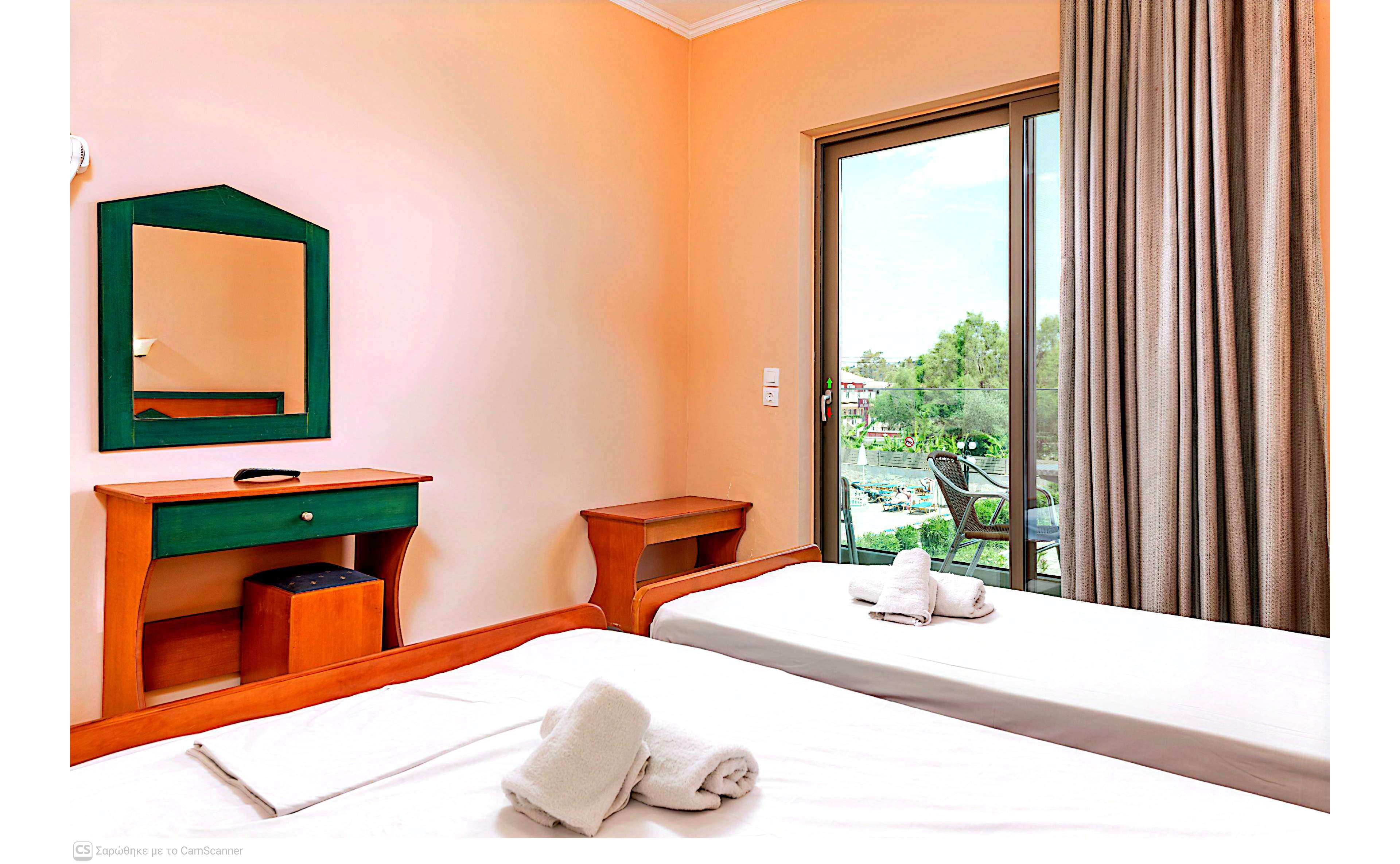 A tidy hotel room features two single beds with neatly folded white towels placed on top. A wooden desk with a mirror and a small stool sits against a peach-colored wall. Floor-to-ceiling curtains frame a sliding glass door that opens to a balcony overlooking greenery and outdoor seating. The space appears clean, bright, and inviting, with natural light enhancing the warm tones of the interior.