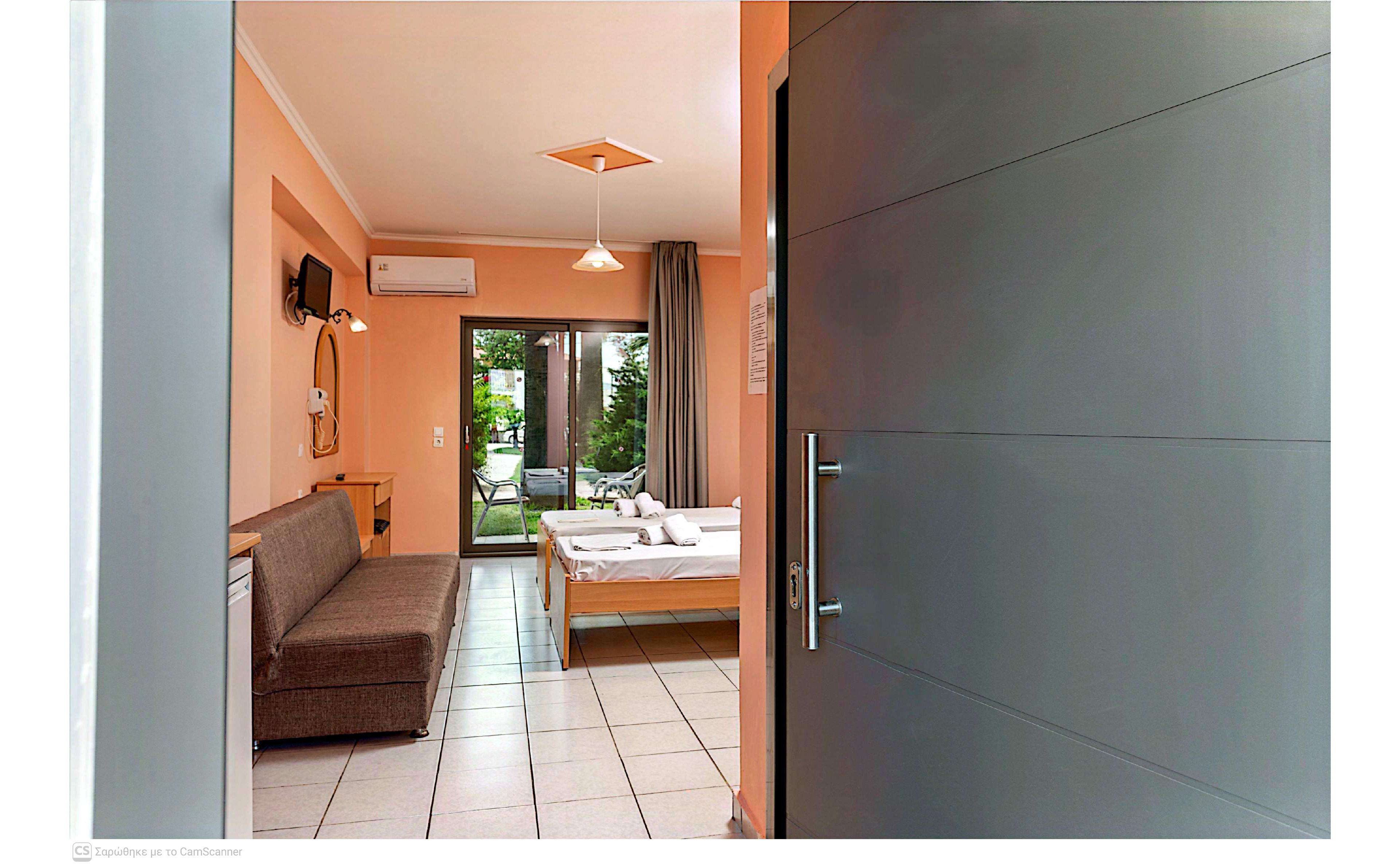 Interior view of a small hotel room seen from the open doorway. The space features a double bed with white linens, a brown sofa, tiled floor, and peach-colored walls. A wall-mounted television and air conditioner are visible above a compact desk area. Glass doors at the far end open to a green garden patio, allowing natural light into the room.
