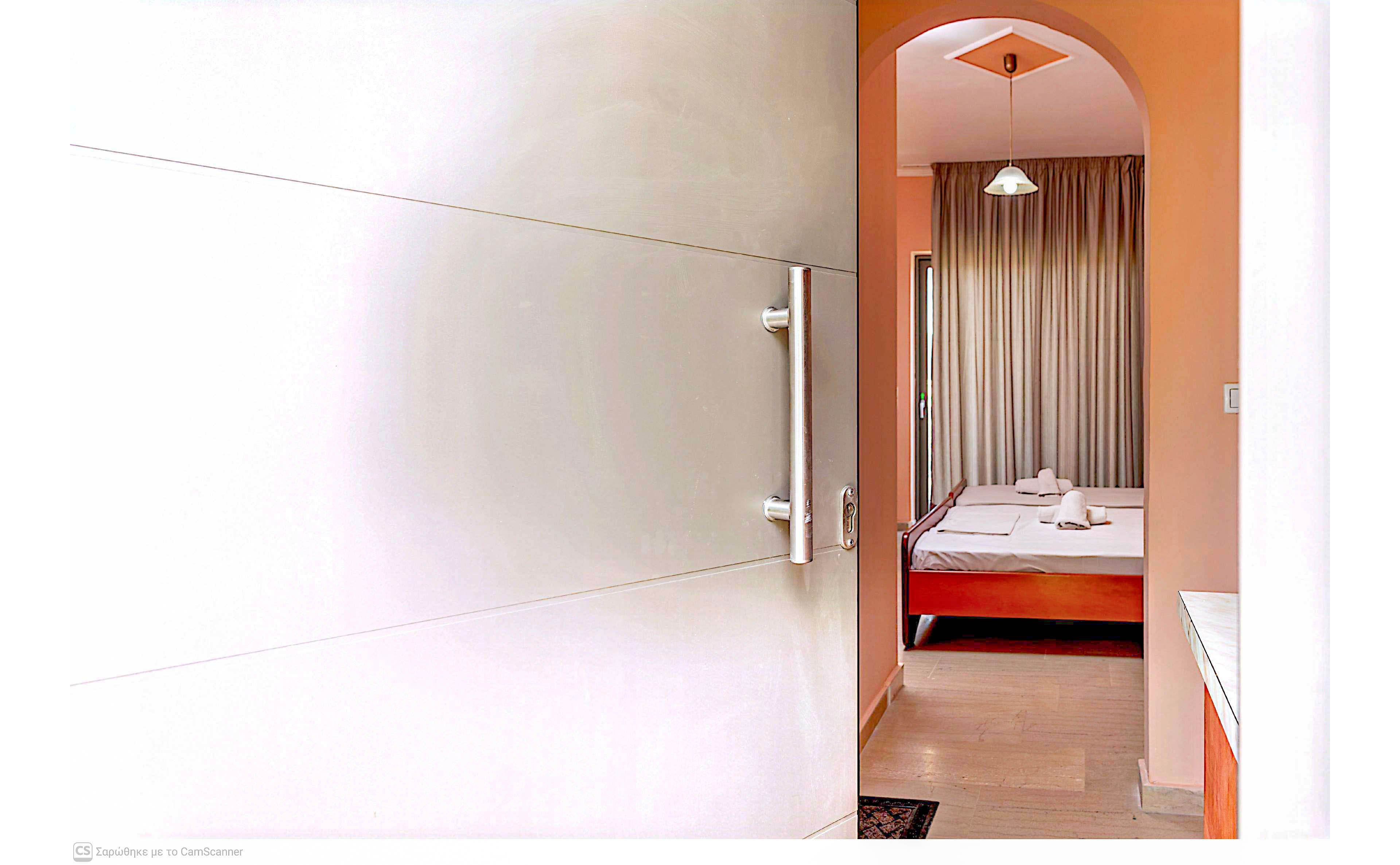 A partially open modern door leads into a neat bedroom. Inside the room, a double bed with white linens and folded towels is visible against a warm-colored wall. Floor-to-ceiling curtains cover the window, and a ceiling light hangs from the arched doorway. The scene suggests a clean, welcoming accommodation interior with simple contemporary decor.