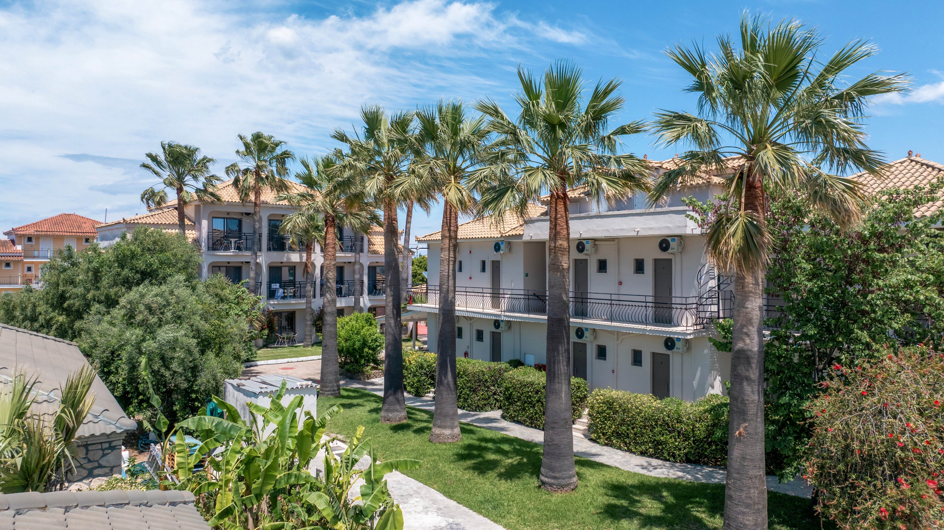 A modern apartment complex is surrounded by tall palm trees and lush greenery under a bright blue sky. The buildings feature balconies with railings and outdoor air conditioning units. A well-kept lawn and garden beds with tropical plants create a resort-like atmosphere. The scene suggests a warm, coastal or Mediterranean setting ideal for vacation or relaxed living.