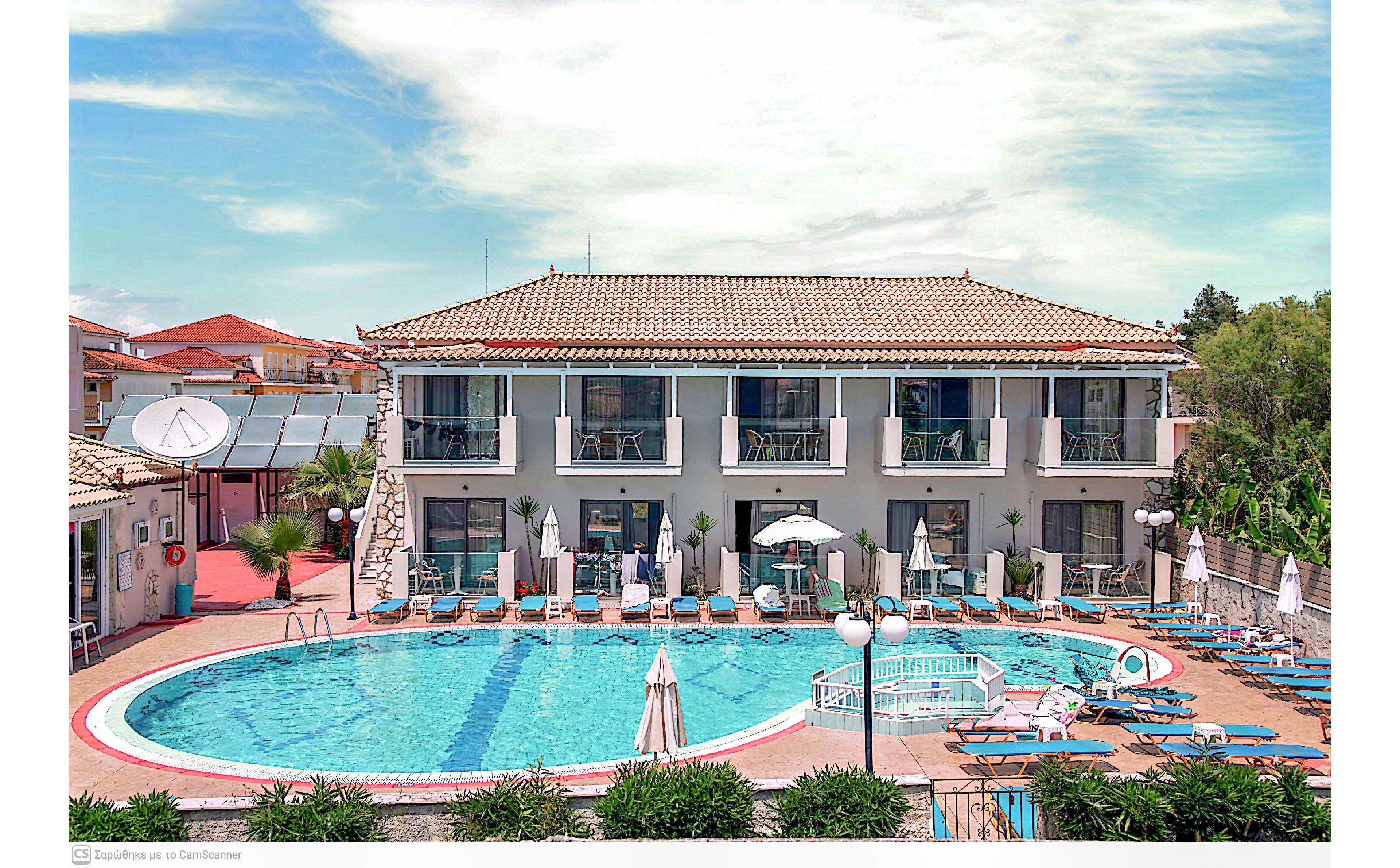 A two-story hotel building overlooks a large curved outdoor swimming pool. Sun loungers and umbrellas are neatly arranged around the poolside terrace, suggesting a relaxing vacation setting. The sky is bright with scattered clouds, and surrounding greenery and nearby rooftops frame the scene. The overall atmosphere is calm and inviting, ideal for summer holidays and leisure travel.