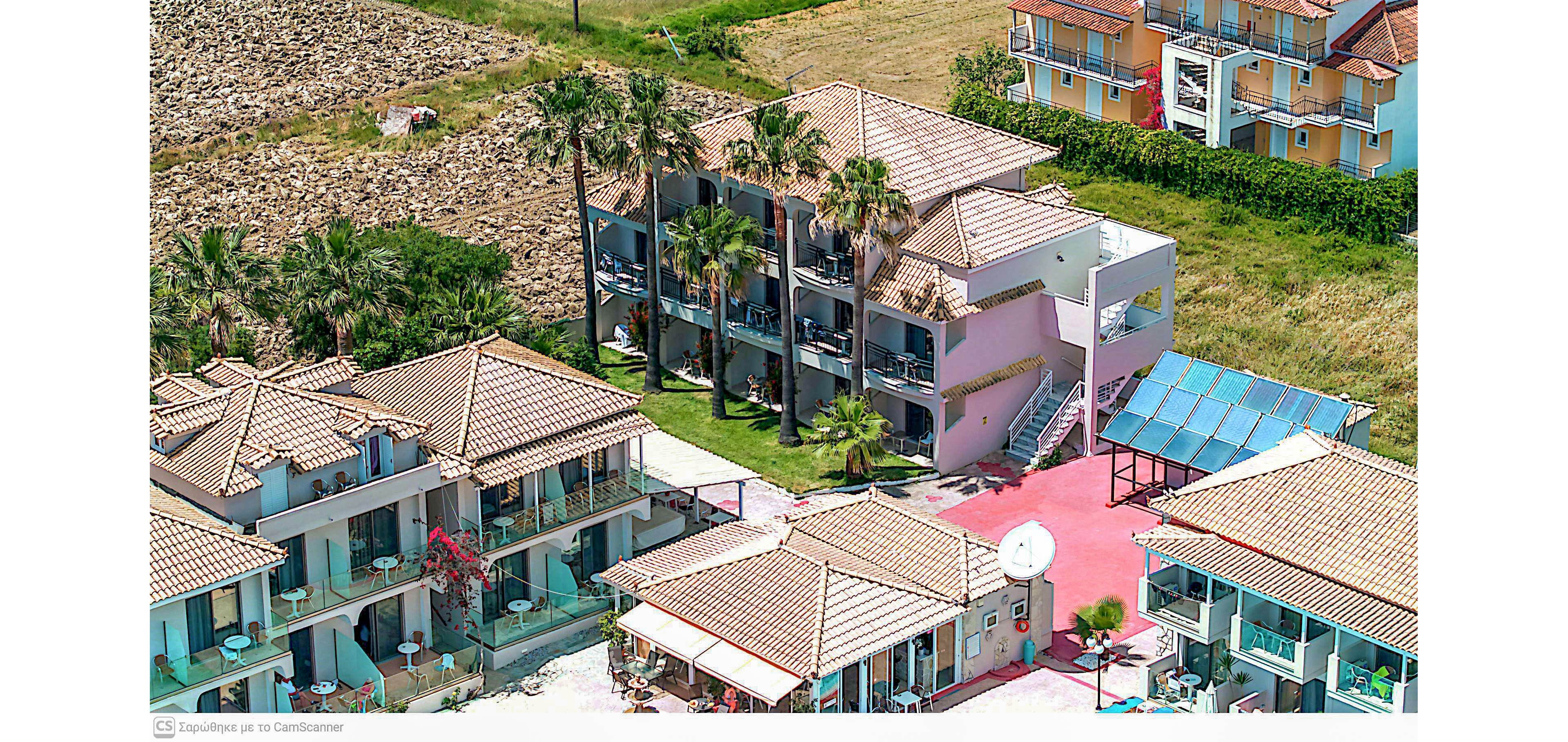 A cluster of low-rise holiday apartment buildings with terracotta roofs is shown from an aerial perspective. The complex features balconies, palm trees, and a central courtyard area with bright red paving. A small pool or covered terrace with a blue roof is visible near one of the buildings. Surrounding the property are fields and additional colorful residential structures, suggesting a coastal or rural Mediterranean resort setting.