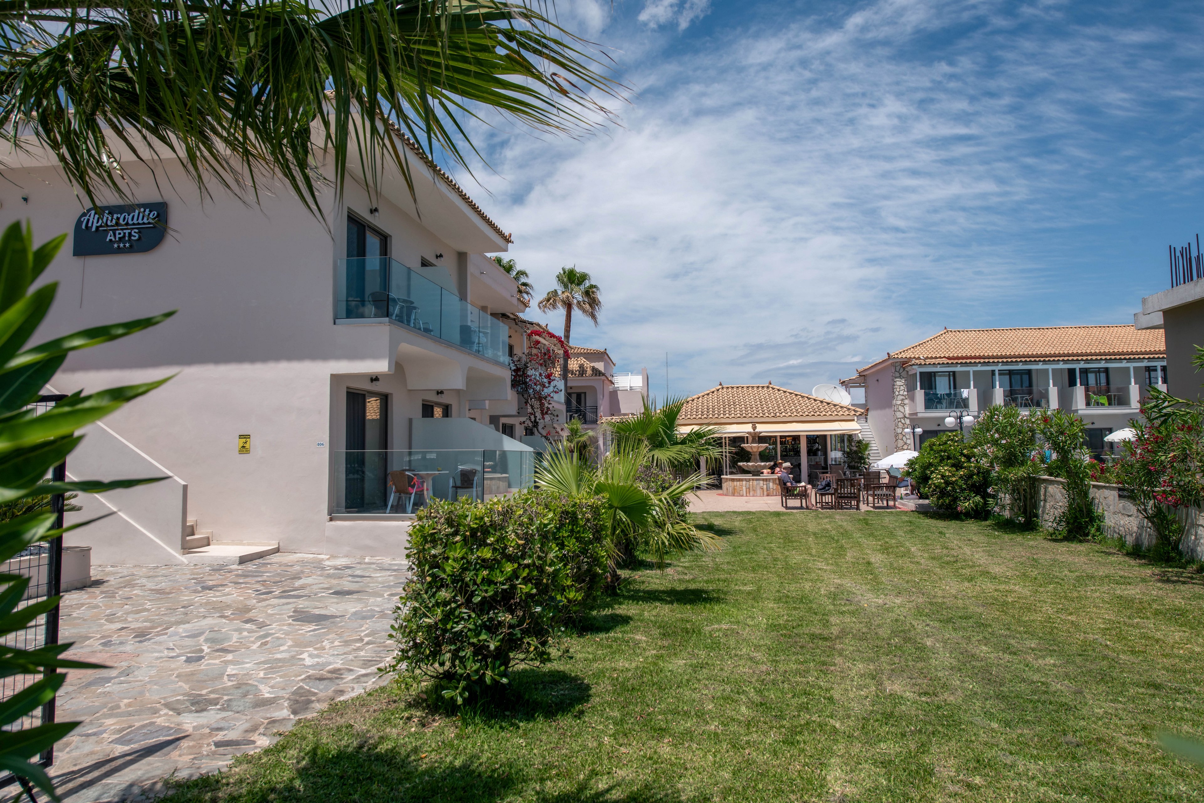 A modern hotel complex with white buildings surrounds a green courtyard lawn under a bright blue sky. Glass balconies and stone walkways lead toward an outdoor terrace area with tables and umbrellas. Palm trees, shrubs, and flowering plants add lush greenery to the scene. The overall atmosphere is relaxed and inviting, suggesting a warm holiday destination.