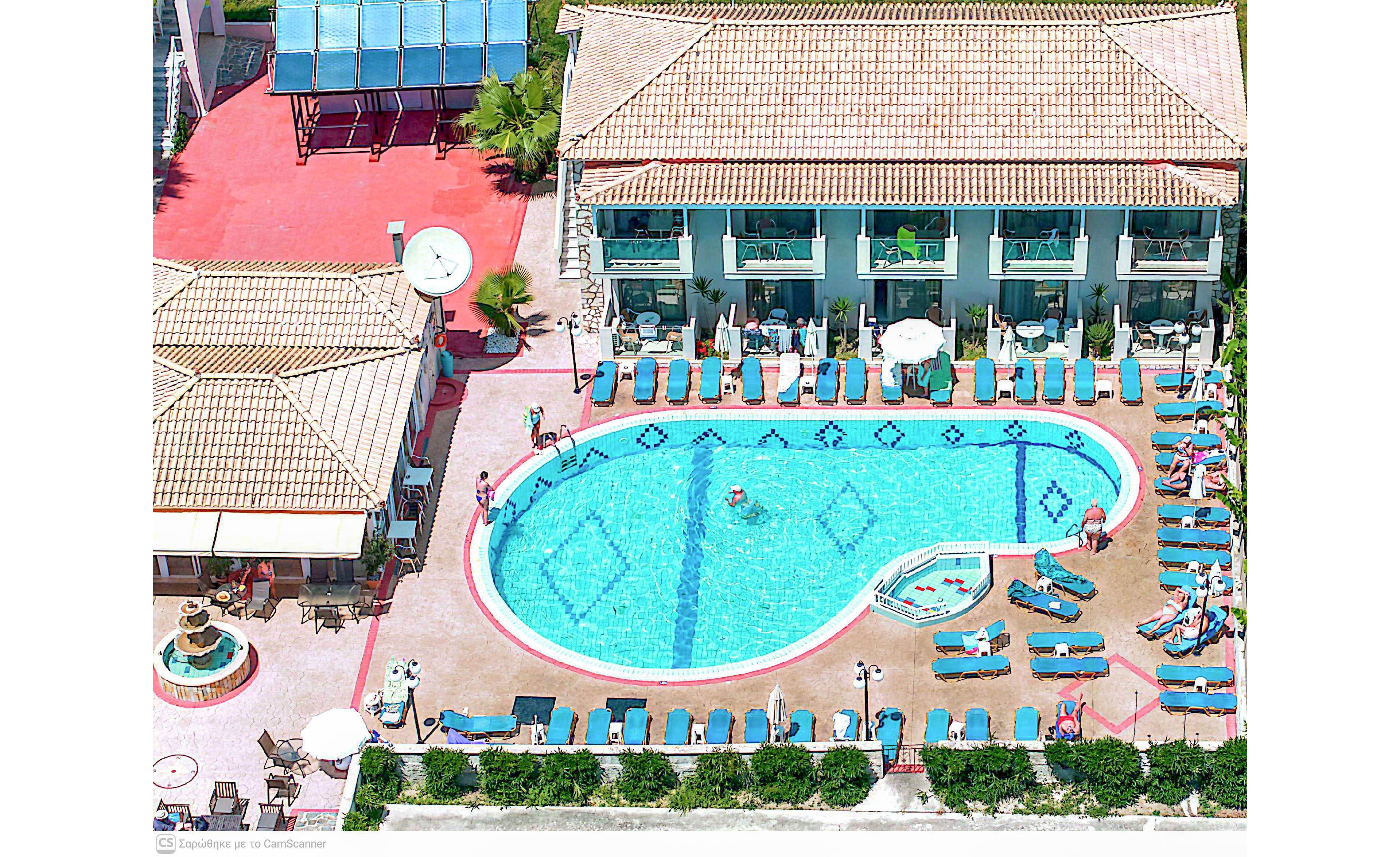 A large outdoor swimming pool is seen from above at a sunny resort. The pool is surrounded by rows of turquoise lounge chairs and umbrellas, with several people relaxing and swimming. Two main buildings with tiled roofs and balconies frame the pool area, suggesting hotel accommodations. Bright blue water, red walkways, and green plants create a vibrant, summery atmosphere.