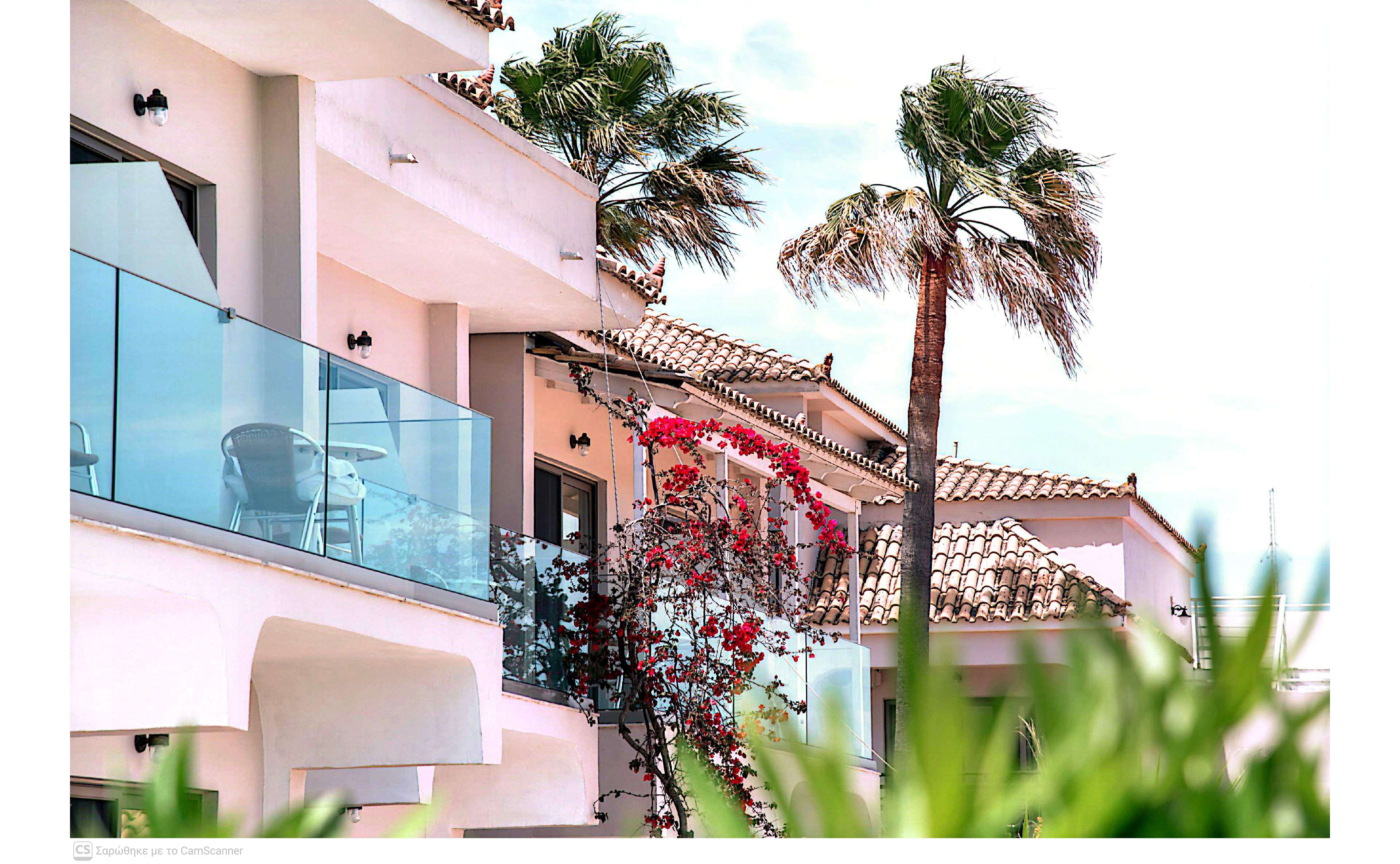 Modern white apartments with tiled roofs stand under a bright, sunny sky. Glass balconies and clean architectural lines give the buildings a contemporary Mediterranean feel. Tall palm trees sway beside the structures, adding a tropical touch. Vibrant flowering vines climb the facade, creating a colorful contrast against the pale walls.