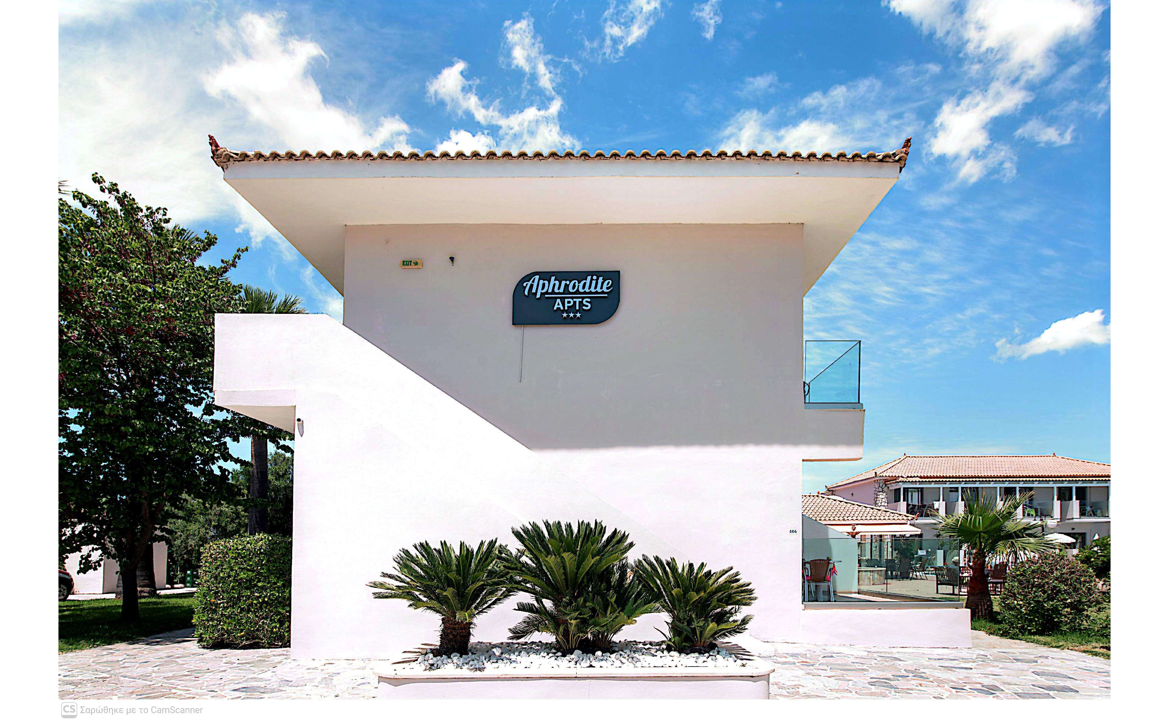 A white modern apartment building labeled "Aphrodite Apts" stands under a bright blue sky with scattered clouds. The structure features clean geometric lines, an exterior staircase, and a small balcony with glass railing. In the foreground, neatly trimmed palm plants and a paved walkway enhance the tidy, resort-like atmosphere. Additional low-rise buildings and greenery are visible in the background, suggesting a holiday complex or residential area.