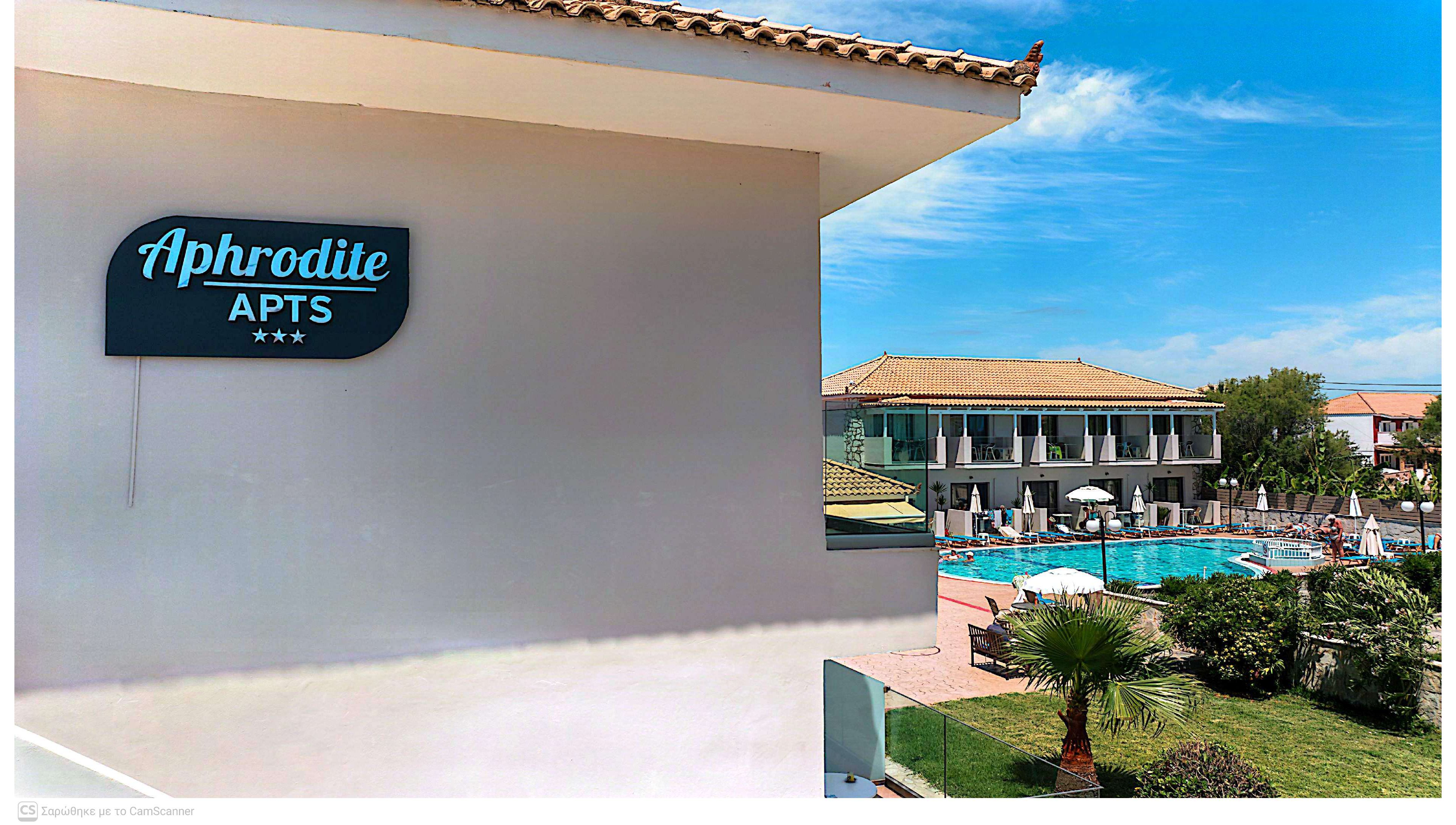 Exterior view of the Aphrodite APTS complex with its sign prominently displayed on a light-colored building. In the background, a large swimming pool is surrounded by sun loungers and umbrellas, with a multi-story accommodation block facing the water. Palm trees and landscaped greenery add a tropical feel to the resort setting. The bright blue sky and clear weather suggest a warm, sunny vacation destination. Three stars are visible beneath the apartment name, indicating the property’s rating.