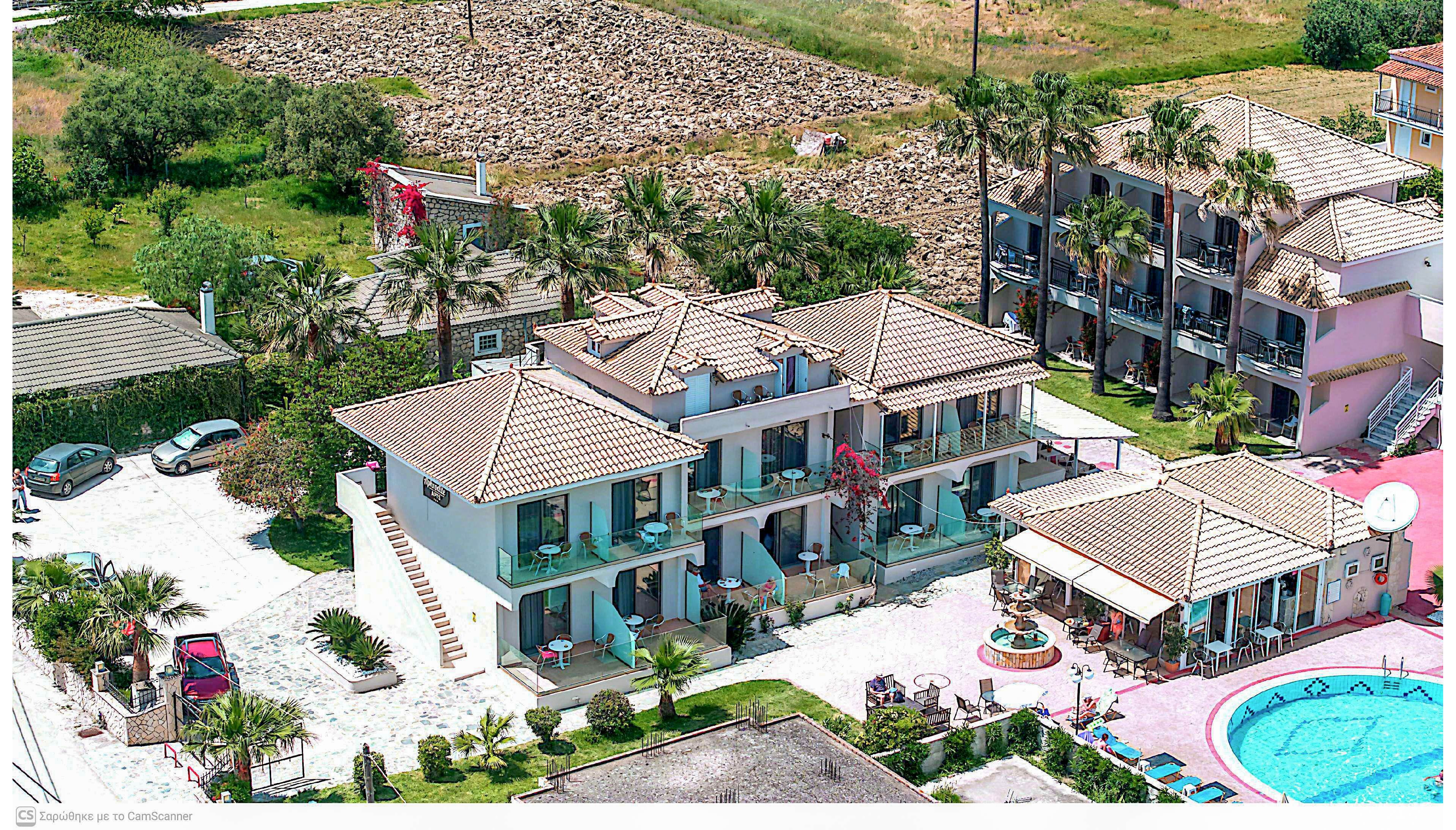 Aerial photograph of a small Mediterranean-style resort with multiple low-rise apartment buildings. The complex features tiled roofs, balconies with outdoor seating, and surrounding palm trees. A bright blue swimming pool with sun loungers and umbrellas is visible in the foreground. The property is bordered by greenery and an open field, suggesting a quiet vacation setting.