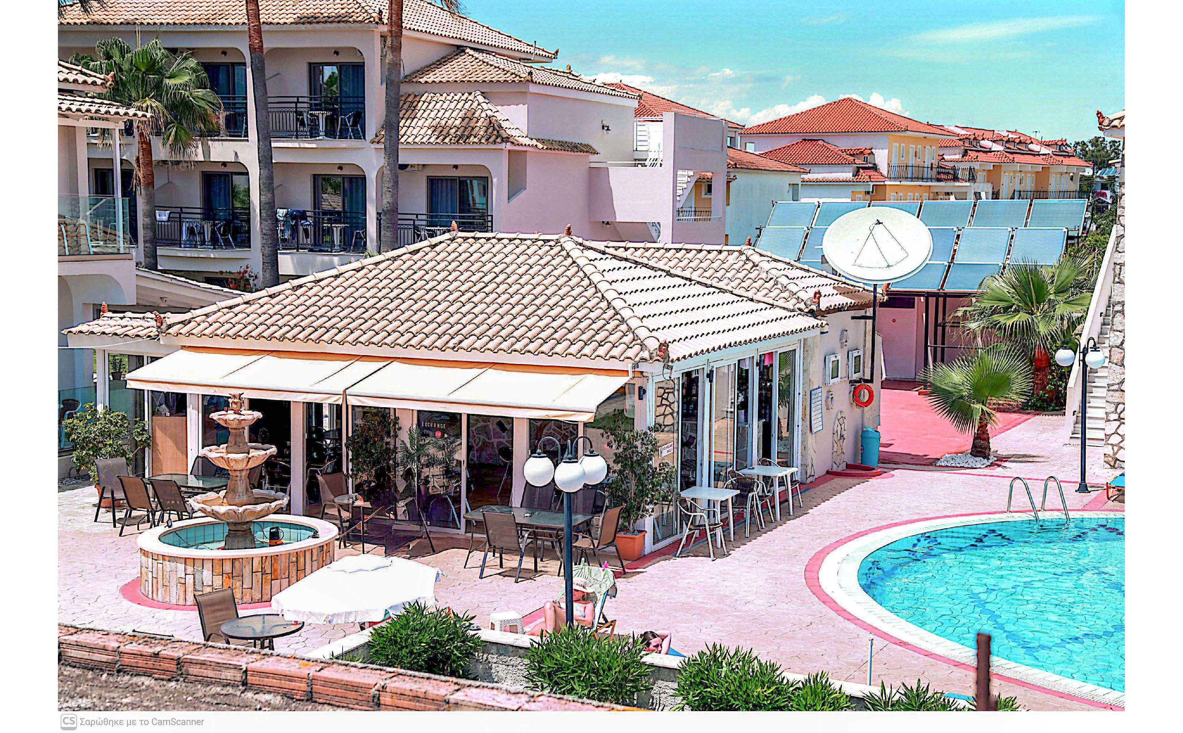An outdoor resort area features a poolside cafe with a tiled roof and surrounding apartment buildings. A clear blue swimming pool curves along the right side, bordered by pink paving and lounge chairs. A decorative fountain and shaded seating area create a relaxed vacation atmosphere. Solar panels and a large satellite dish are visible on the roof, suggesting modern amenities in a sunny coastal setting.
