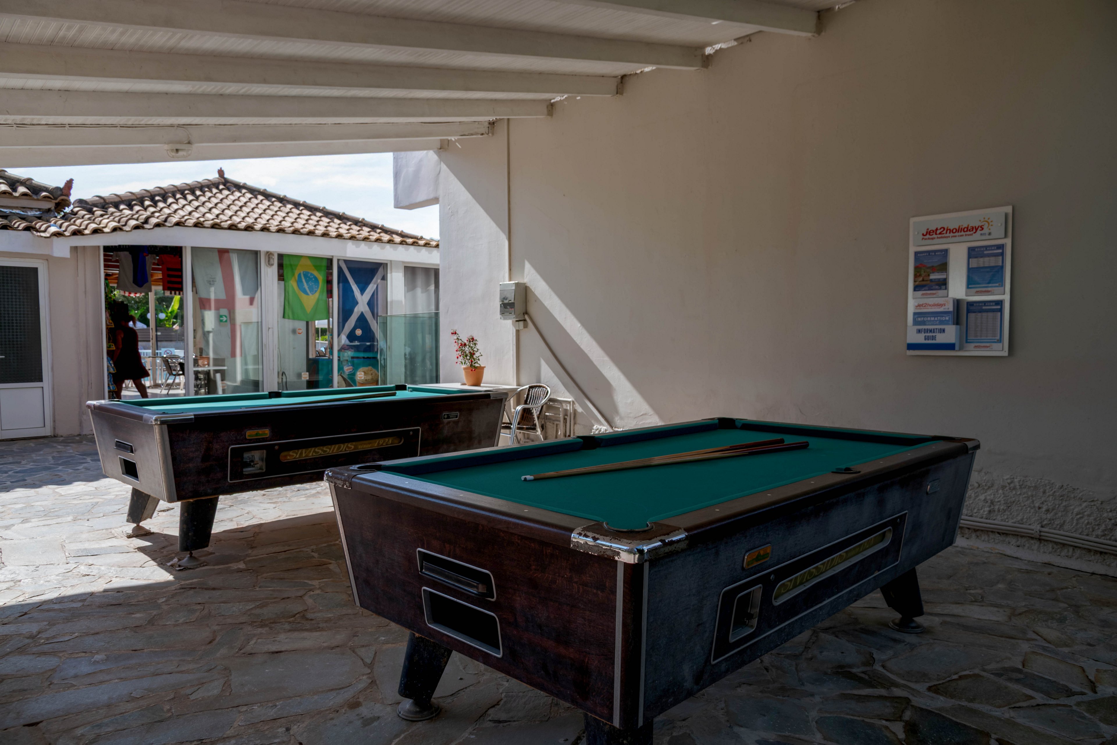 Two pool tables stand in a shaded, open-air area with a stone floor. Sunlight streams in from the side, casting strong shadows across the tables and ground. In the background, a building with tiled roof and glass doors displays colorful flags inside. A wall-mounted notice board with printed information is visible on the right side of the image.