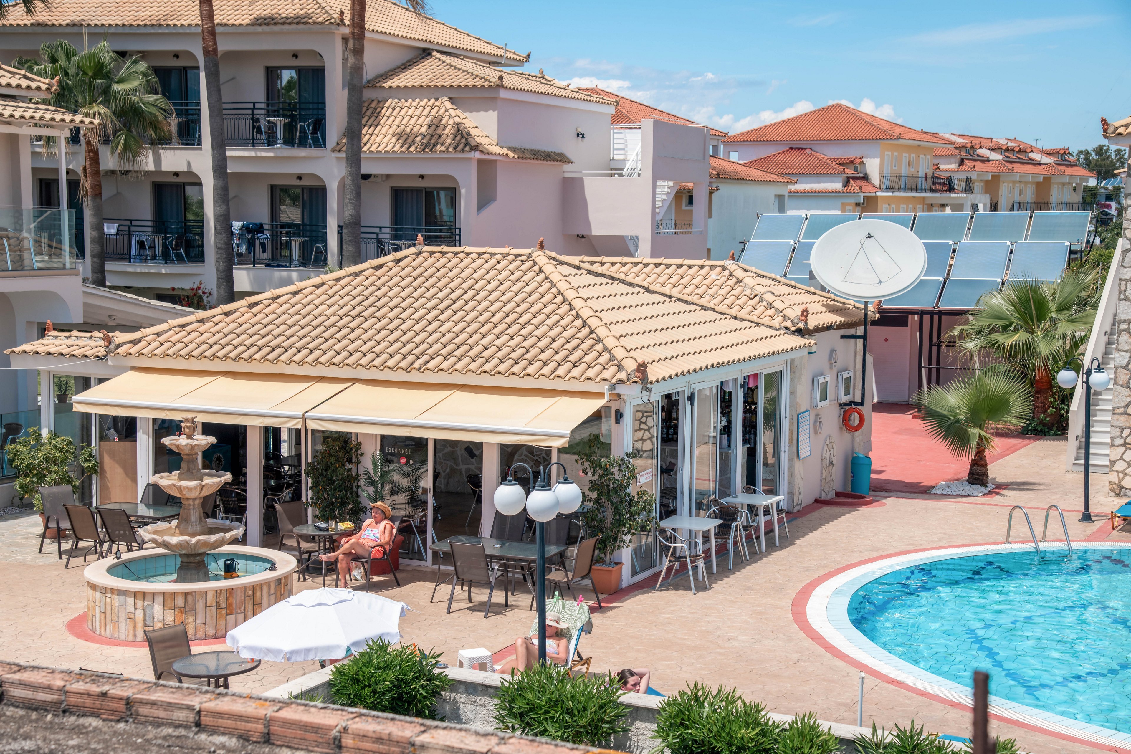 A Mediterranean-style hotel complex features a poolside cafe with a tiled roof and outdoor seating. Guests relax at tables near a decorative multi-tier fountain in the courtyard. A curved swimming pool with clear blue water occupies the foreground, surrounded by sun loungers and palm trees. In the background, apartment-style hotel buildings and rooftop solar panels complete the sunny resort setting.