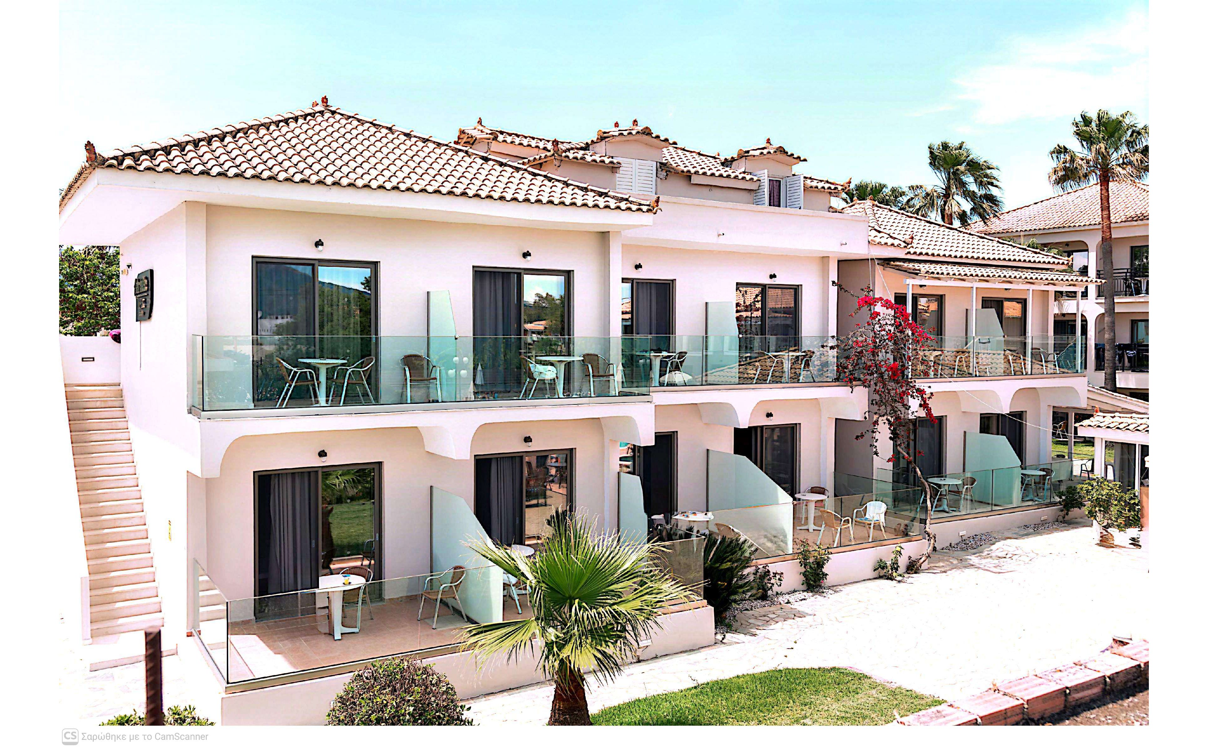 A two-story Mediterranean-style resort building features multiple guest apartments with glass-railed balconies and patios. Outdoor chairs and tables are arranged on each balcony, suggesting a relaxing holiday setting. The light-colored facade contrasts with the blue-tinted glass and dark window curtains. Palm trees, flowering plants, and a sandy pathway enhance the sunny, coastal atmosphere.