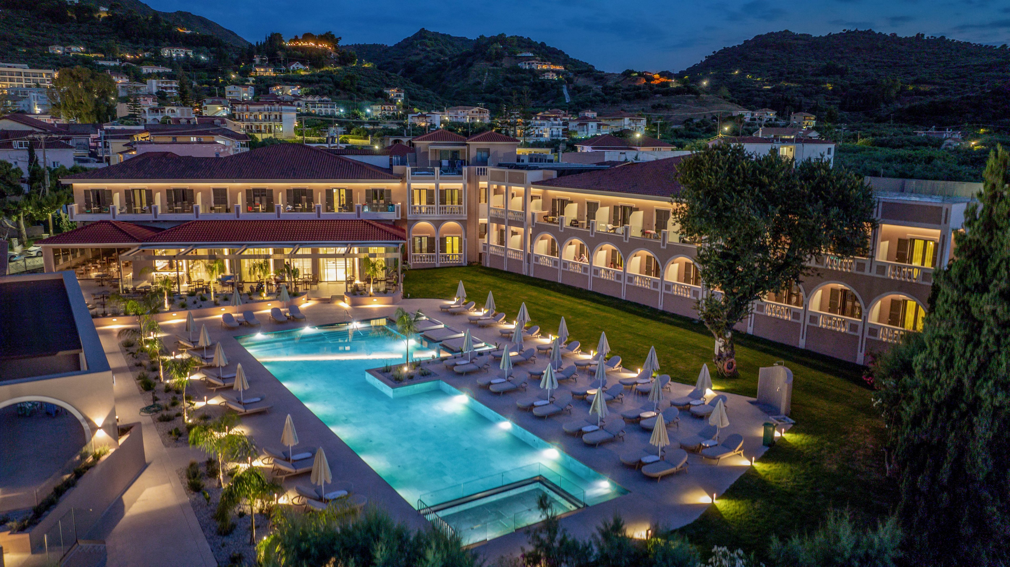A large upscale resort complex is shown at dusk with an expansive central swimming pool glowing in blue light. Rows of sun loungers and closed umbrellas surround the pool, suggesting a tranquil, end-of-day atmosphere. The multi-story buildings feature warm interior lighting, balconies, and arched walkways overlooking the courtyard. In the background, hills and scattered houses frame the property, enhancing the scenic vacation setting.