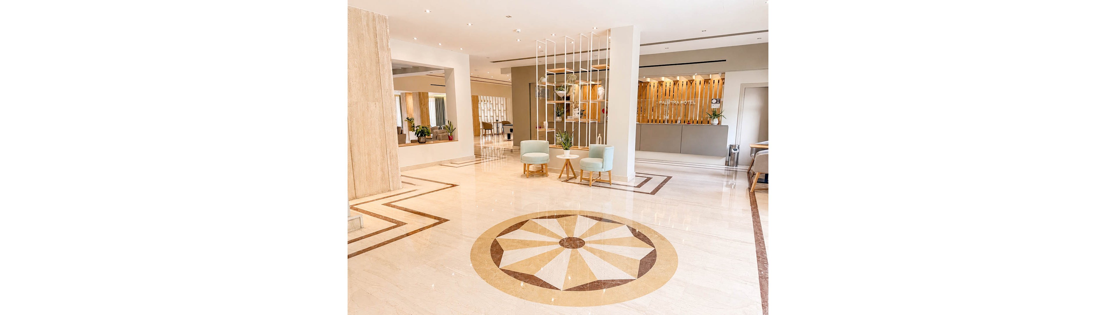 Spacious hotel lobby interior featuring a polished marble floor with a circular geometric inlay. Two pastel armchairs and a small wooden table create a cozy seating area near a decorative white metal partition. The reception desk with vertical wooden slats is visible in the background, adding warmth and texture. Large windows and neutral tones give the space an airy, contemporary feel.