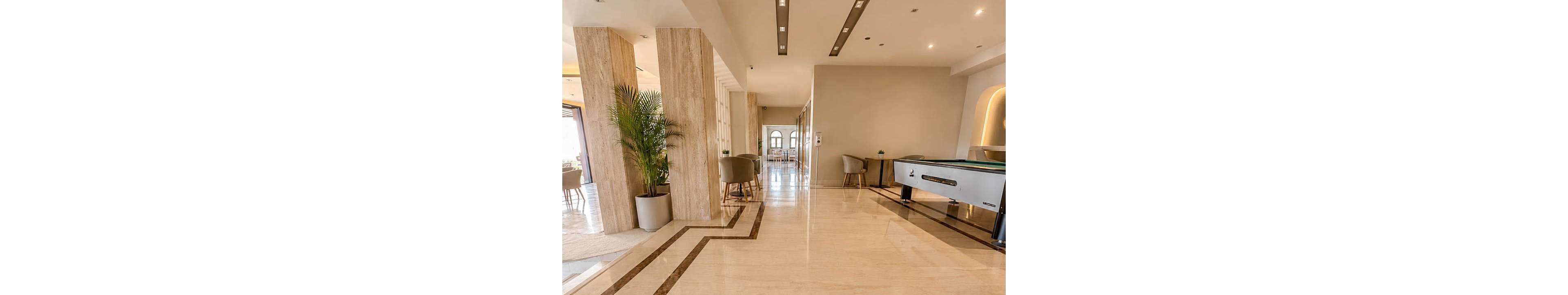 A spacious, contemporary lobby features polished marble floors with geometric brown inlays. Tall stone columns and recessed ceiling lights create a clean, upscale atmosphere. Lounge chairs and small tables line the corridor, while a foosball table adds a casual entertainment element. Natural light filters in from the far doorway, enhancing the warm neutral color palette.