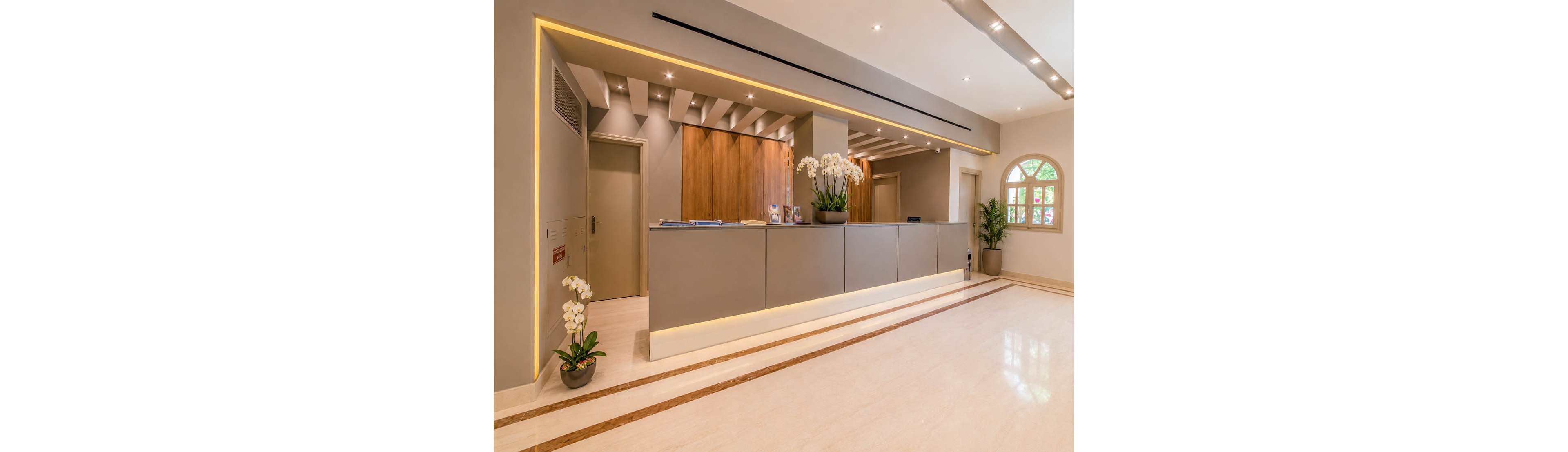 A contemporary reception area features a long, minimalist desk with grey panels and warm wooden accents. The lobby is brightly lit with recessed ceiling lights and subtle linear lighting along the walls. Potted orchids and green plants add a touch of nature to the clean, polished floor and neutral color palette. A window with an arched frame and closed doors complete the calm, professional interior setting.