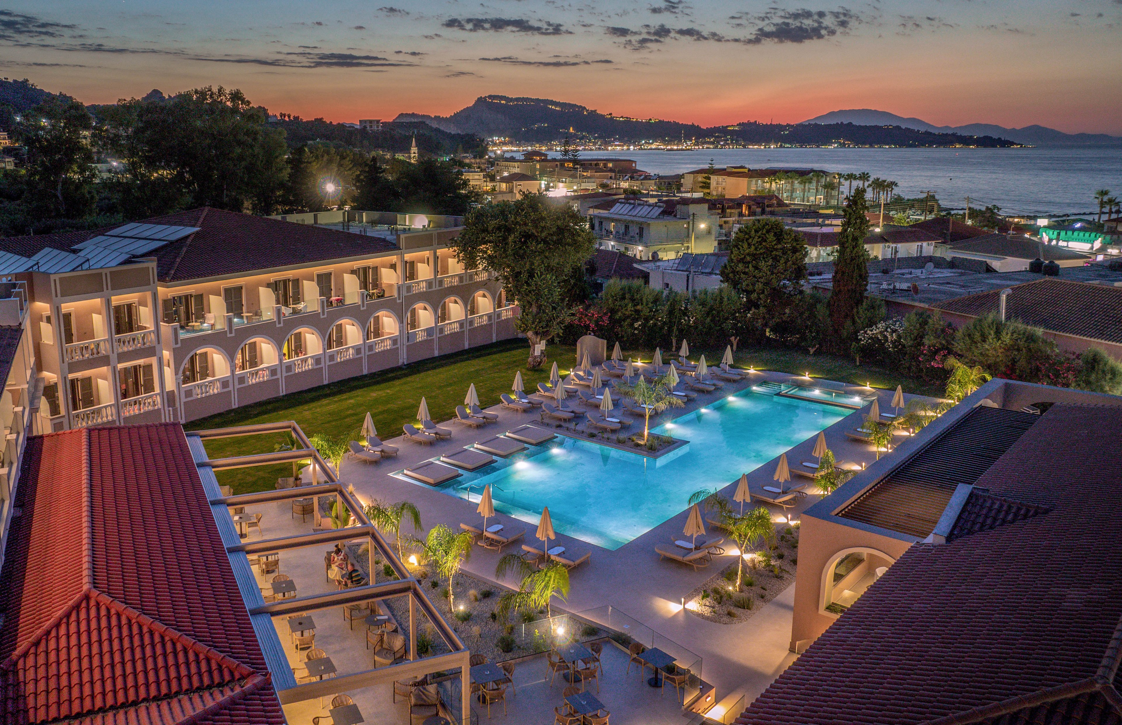 An elevated view shows a luxury resort with a large illuminated swimming pool surrounded by sun loungers and umbrellas. The hotel buildings feature arched balconies and red-tiled roofs, framing the central courtyard area. Beyond the resort, a coastal town and calm bay are visible under a colorful evening sky with soft clouds. The overall scene conveys a tranquil vacation atmosphere with warm lighting and scenic waterfront surroundings.