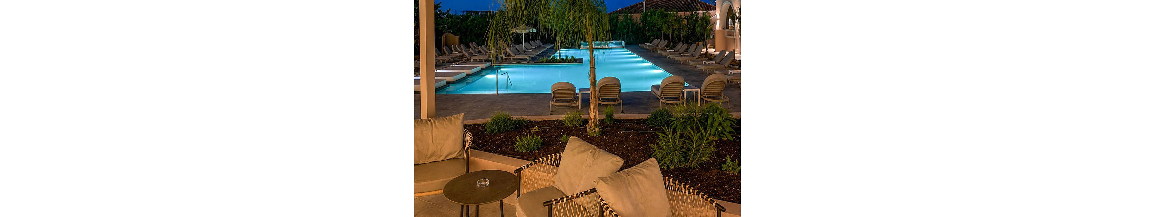 An upscale outdoor lounge area overlooks a long illuminated swimming pool at dusk. Comfortable cushioned chairs and small tables sit under a covered patio in the foreground. The pool is lined with sun loungers and surrounded by greenery and low buildings with warm lights. The deep blue evening sky and soft pool lighting create a calm, inviting atmosphere.