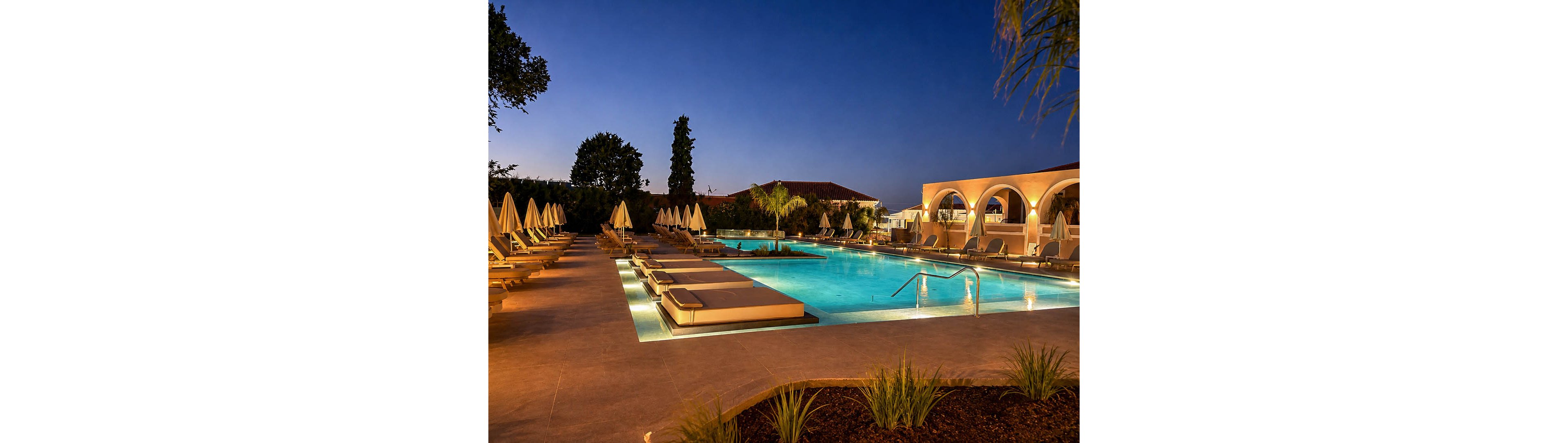 A large illuminated swimming pool is shown at dusk in an upscale resort setting. Lounge chairs with cushions and closed umbrellas line both sides of the pool, creating a symmetrical, inviting scene. Warm architectural lighting highlights arched structures and surrounding buildings against the deep blue evening sky. Tropical plants and neatly landscaped greenery frame the foreground, enhancing the tranquil vacation atmosphere.