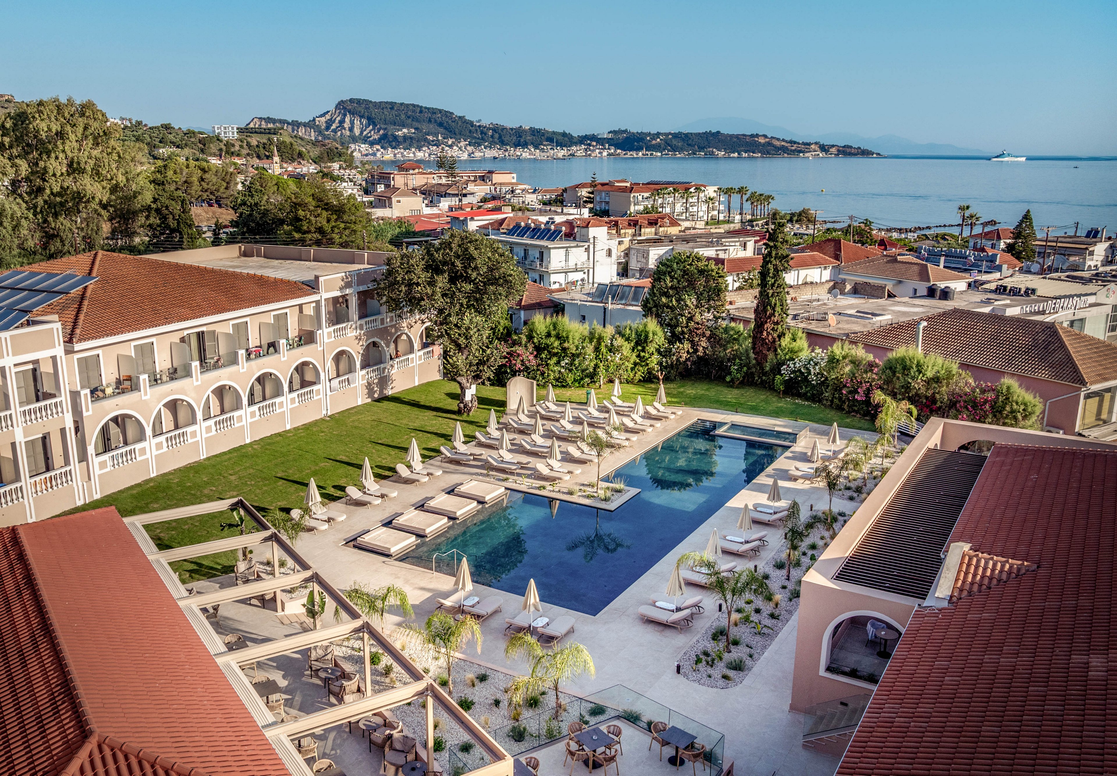 Aerial view of a Mediterranean-style resort featuring a large rectangular swimming pool surrounded by sun loungers and umbrellas. The hotel buildings with arched balconies frame the central courtyard, while landscaped gardens and palm trees add greenery. Outdoor seating and shaded pergolas create inviting social areas near the pool. In the background, a coastal town and calm sea with distant hills complete the scenic vacation setting.