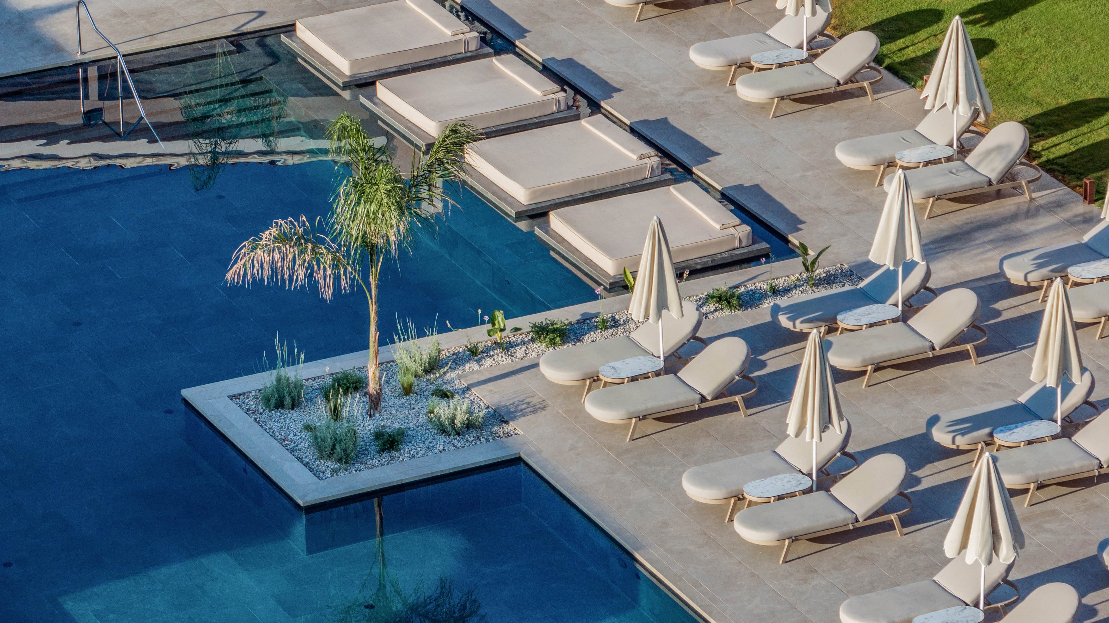 A contemporary outdoor swimming pool is lined with cushioned sun loungers and closed umbrellas, creating a serene resort atmosphere. The pool features clean geometric shapes, steps leading into the water, and an elevated platform with additional daybeds. A small landscaped island with a palm tree and white stones adds a decorative focal point beside the water. The scene is empty of people, emphasizing relaxation, luxury, and calm in a sunny setting.