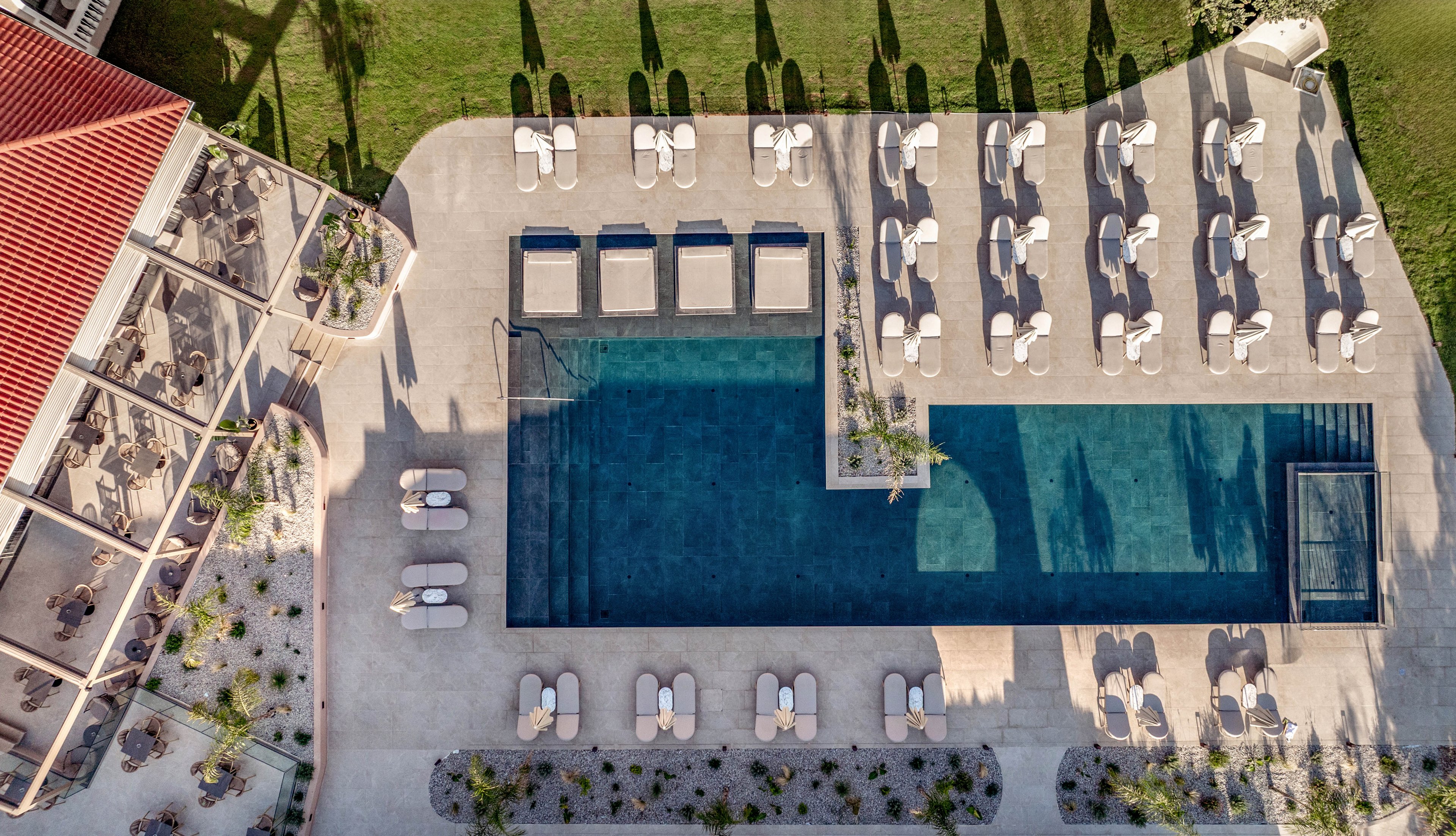 A rectangular outdoor swimming pool is seen from above, surrounded by neatly arranged lounge chairs and umbrellas. The pool area features clean geometric lines, with a smaller adjoining pool or spa section on one side. Adjacent to the deck is a covered terrace with seating, suggesting a resort or hotel setting. Sunlit concrete surfaces, green lawn, and landscaped planters create a bright, inviting atmosphere.