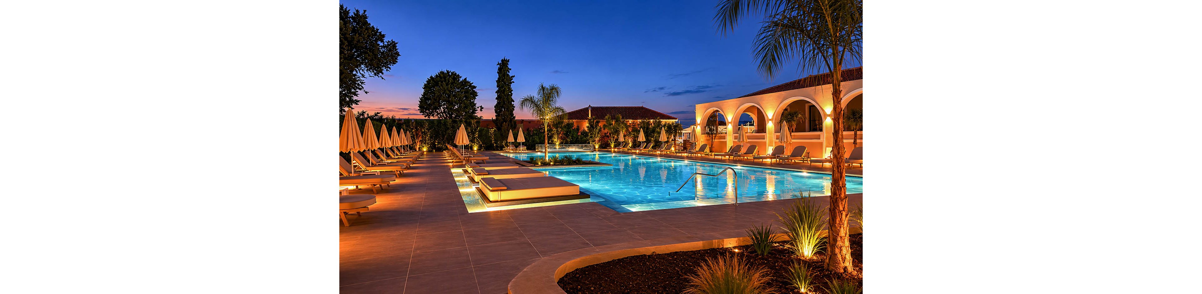 A large illuminated swimming pool is featured at a luxury resort during sunset, surrounded by sun loungers and umbrellas. Warm architectural lighting highlights the arched building and tiled pool deck, creating a tranquil vacation atmosphere. Palm trees and landscaped plants frame the scene, reflecting softly in the calm blue water. The sky transitions from deep blue to warm orange tones, enhancing the serene and inviting mood.