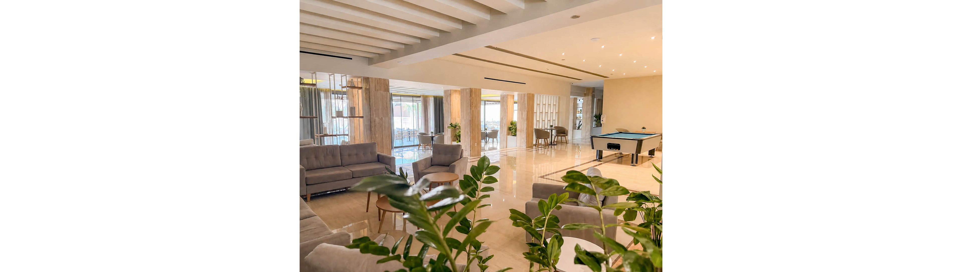 A spacious contemporary lounge area features neutral-toned sofas and armchairs arranged for relaxed seating. Large windows and glass doors allow natural light to fill the polished floor space, with potted green plants adding a touch of nature. In the background, a pool table stands near additional seating, suggesting recreational use in a hotel or resort setting. The overall design is clean and modern, with warm beige surfaces and subtle decorative elements.