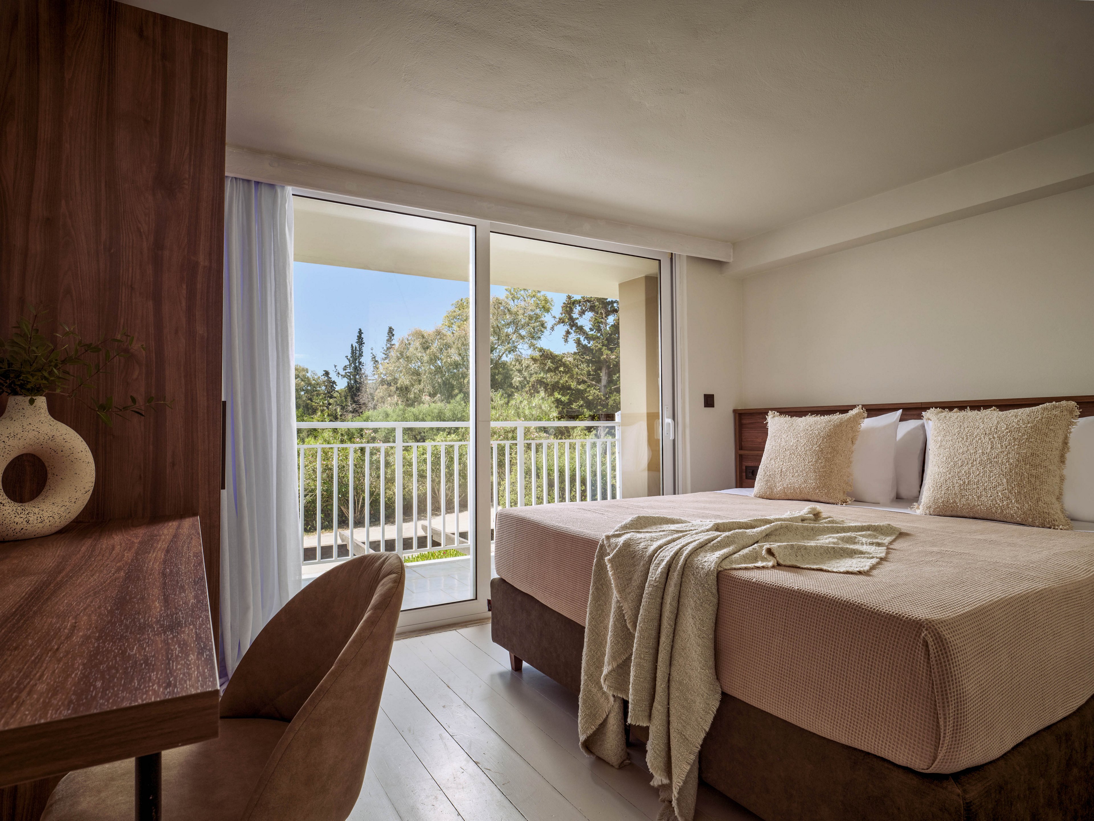 A contemporary hotel bedroom features a neatly made double bed with neutral-toned bedding and textured pillows. Large sliding glass doors open to a balcony with a white railing and a view of trees and greenery outside. A wooden desk and chair sit to the left, adding warmth to the minimalist interior. Soft natural light fills the room, creating a calm and inviting atmosphere.