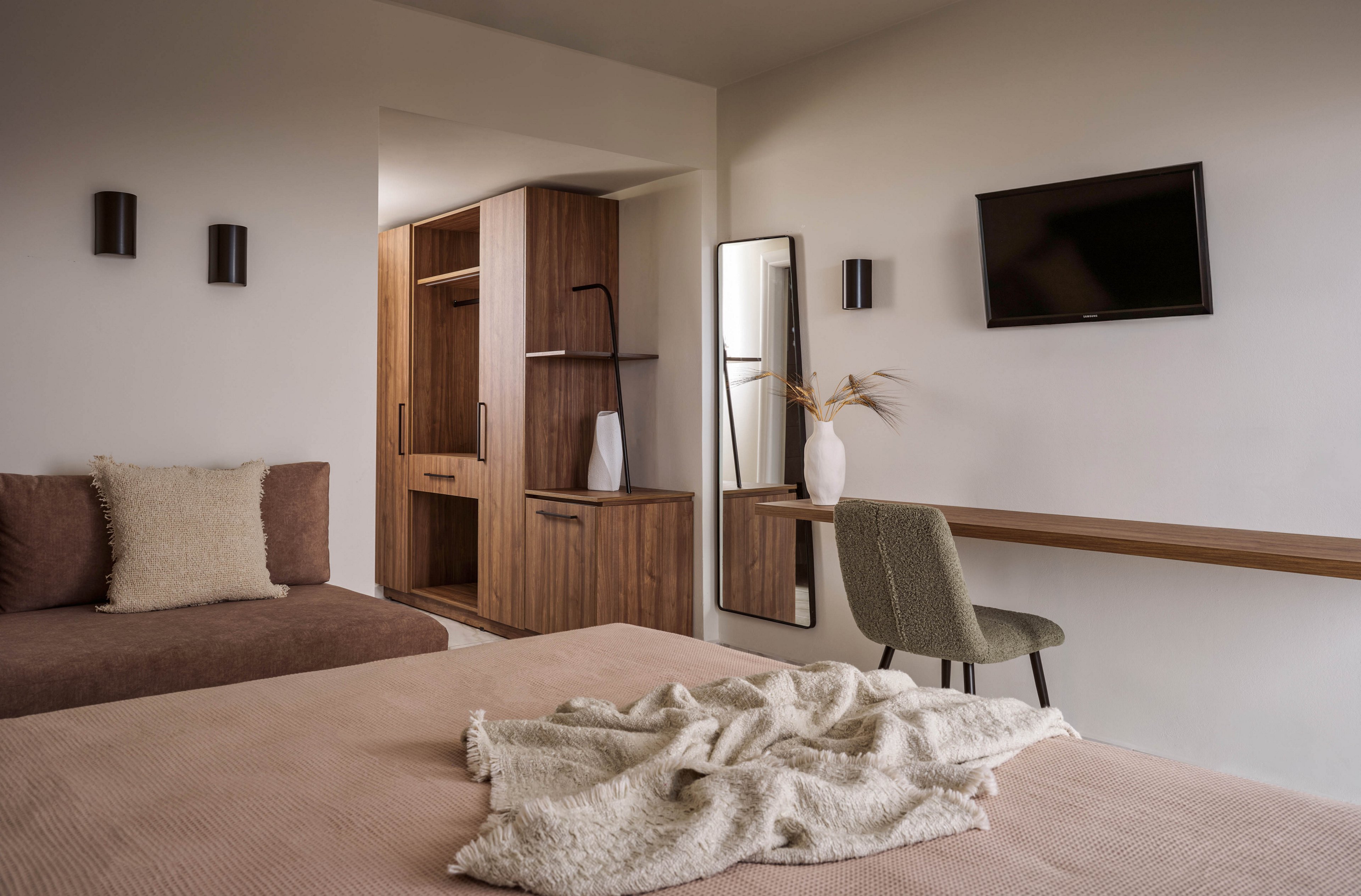 A contemporary bedroom features a neatly made bed with a soft beige blanket and textured cushion. In the background, a built-in wooden wardrobe and shelving unit add warmth and storage. A slim desk with a green upholstered chair sits beneath a wall-mounted television and next to a tall mirror. Neutral tones, clean lines, and simple decor create a calm, minimalist atmosphere.
