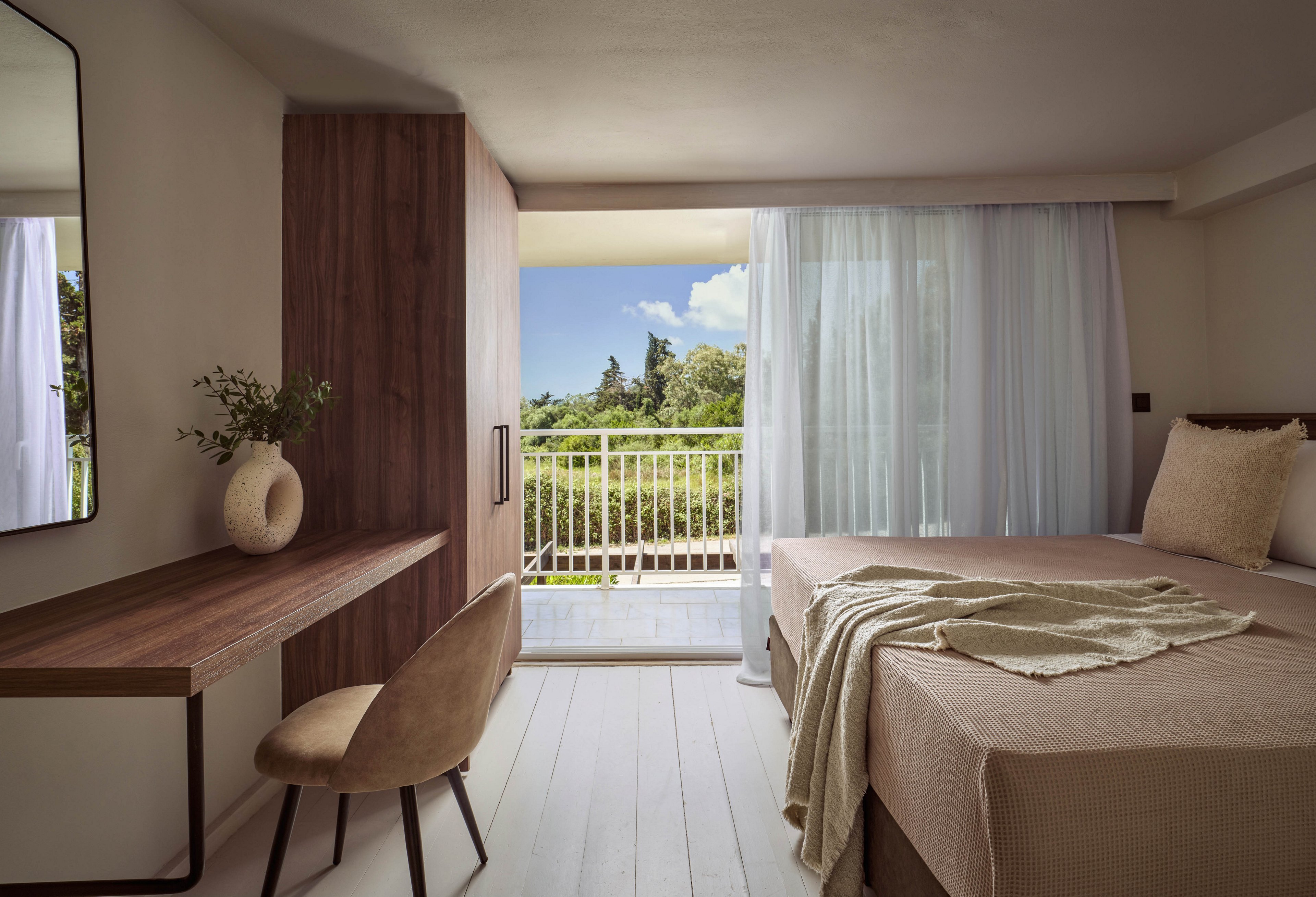 A modern minimalist hotel room features a single bed with neutral bedding and a light throw blanket. A wooden desk with a vase of greenery and a cushioned chair sits opposite the bed. Sheer white curtains frame a large sliding glass door that opens to a balcony. Beyond the balcony, lush green trees and a bright blue sky create a serene outdoor view.