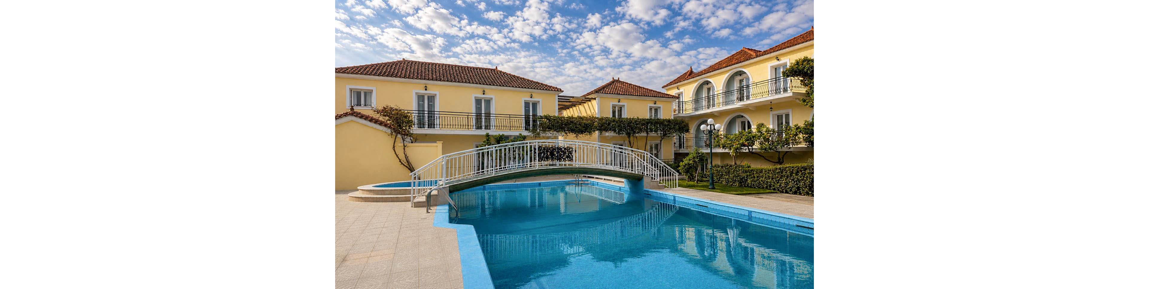 A bright yellow Mediterranean-style villa surrounds a clear blue swimming pool. A white arched footbridge crosses over the pool, connecting the tiled terrace areas. The building features red-tiled roofs, balconies with railings, and neatly trimmed greenery. Soft clouds scatter across the blue sky, enhancing the warm and inviting resort atmosphere.