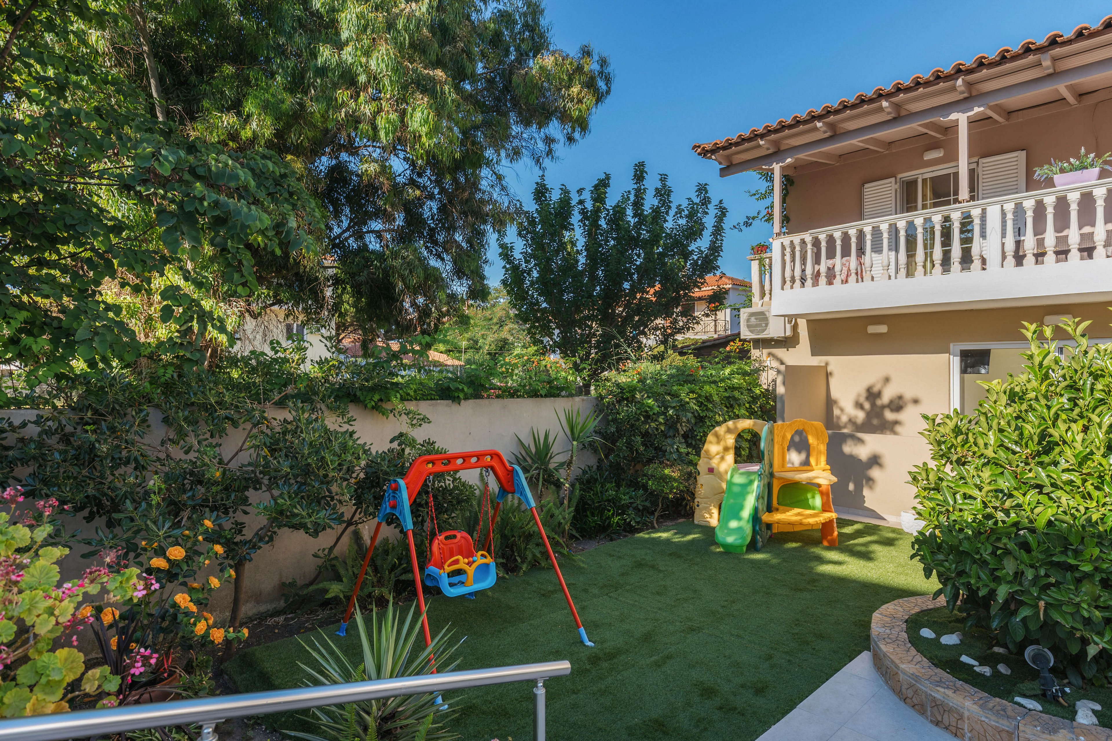 A small backyard garden features a colorful children's playground set on artificial grass. A red and blue swing frame and a yellow plastic playhouse with a green slide stand in the center of the yard. Lush green plants, shrubs, and trees surround the play area, creating a cozy and private outdoor space. In the background, a beige two-story house with a balcony and tiled roof overlooks the garden under a clear blue sky.
