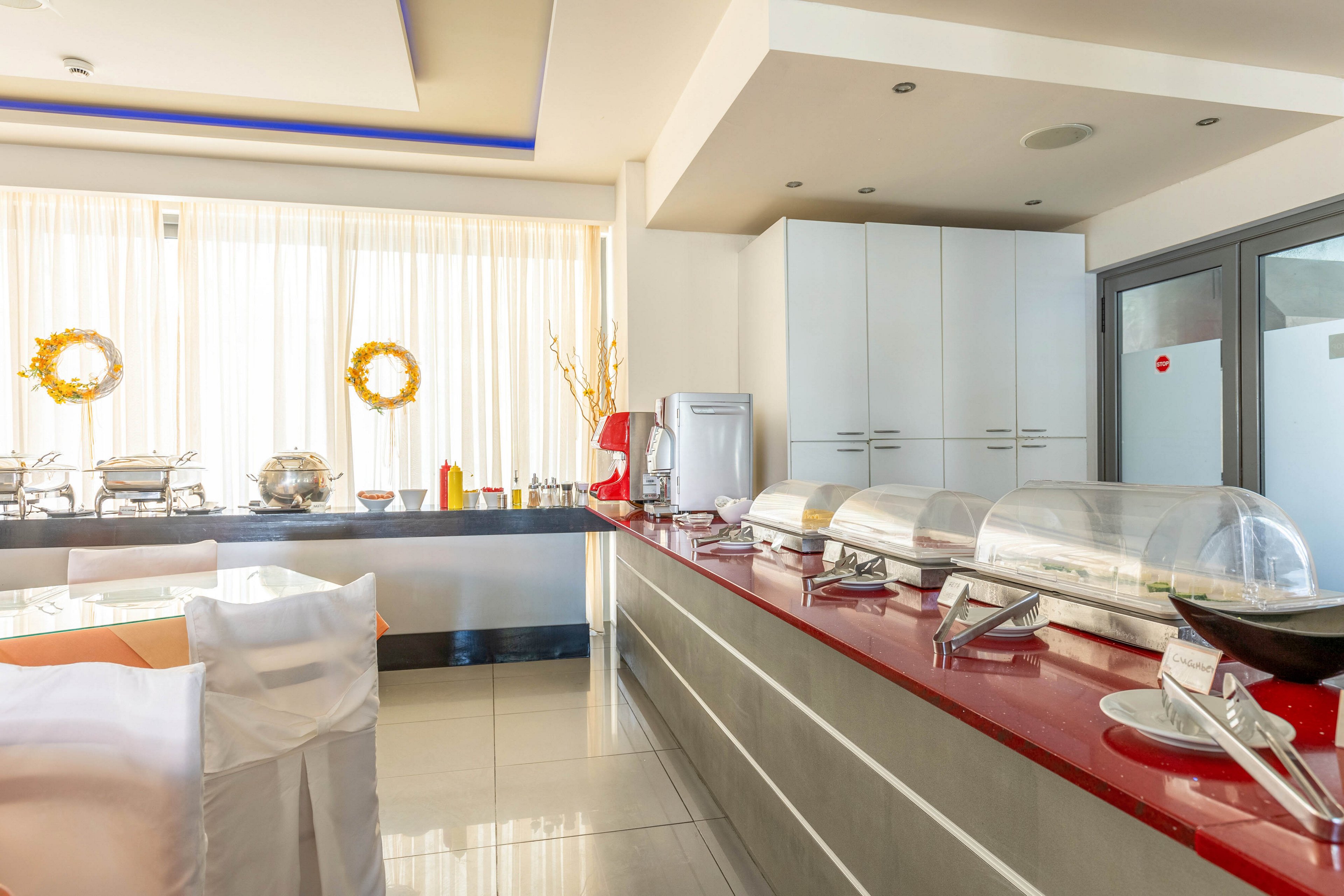A modern buffet area features a long red countertop lined with covered chafing dishes and serving utensils. The space appears to be part of a hotel or restaurant dining room with neatly arranged tables and chairs. Large windows with sheer curtains allow bright daylight to fill the room, highlighting the clean, minimalist design. Decorative wreaths and a tall vase with branches add a subtle festive touch to the otherwise neutral interior.
