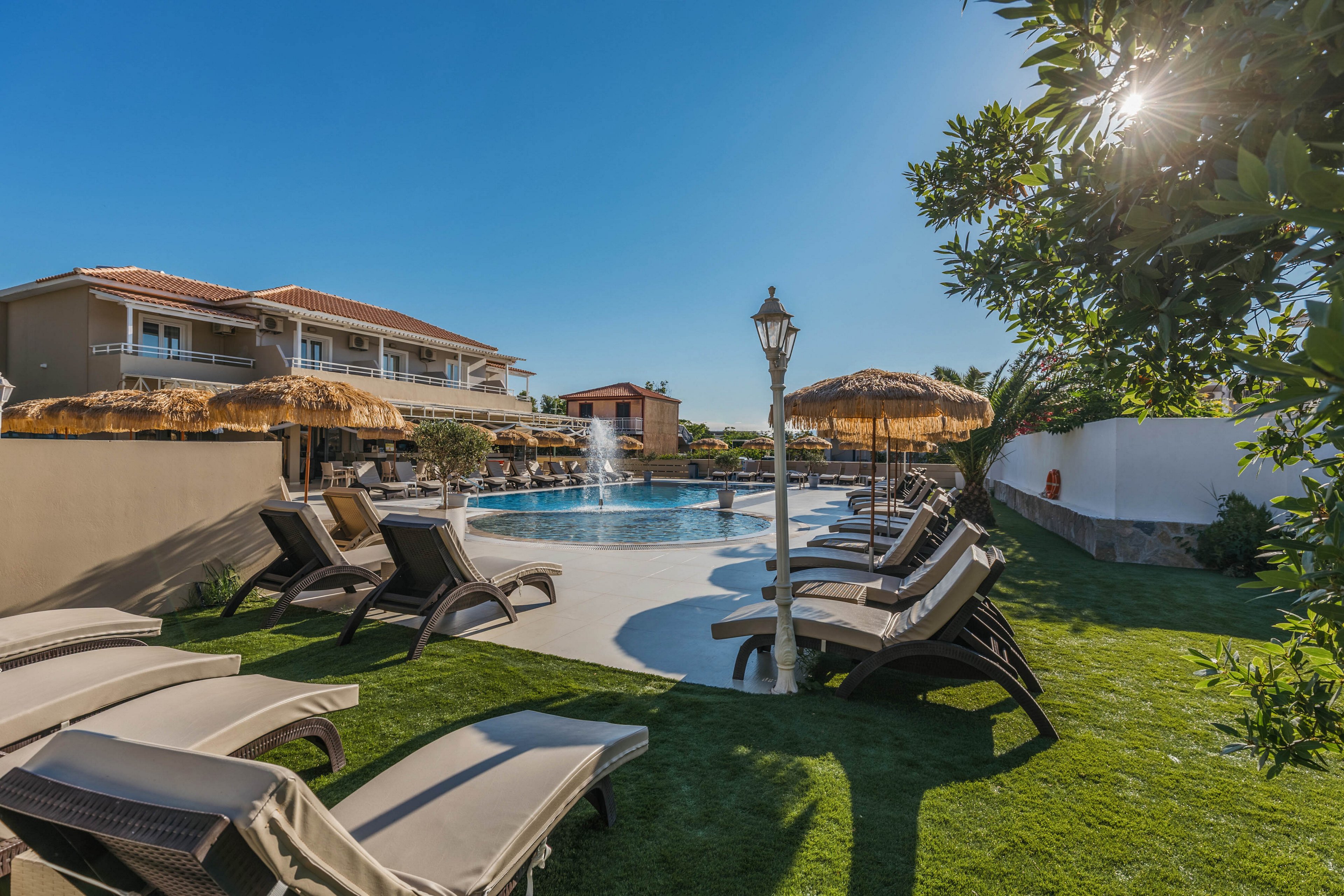 An outdoor resort pool area is shown under a clear blue sky. Multiple lounge chairs line the poolside, shaded by straw parasols on a neatly maintained lawn. In the background, low-rise hotel buildings with balconies overlook the water. The bright daylight and calm setting create a relaxed vacation atmosphere.