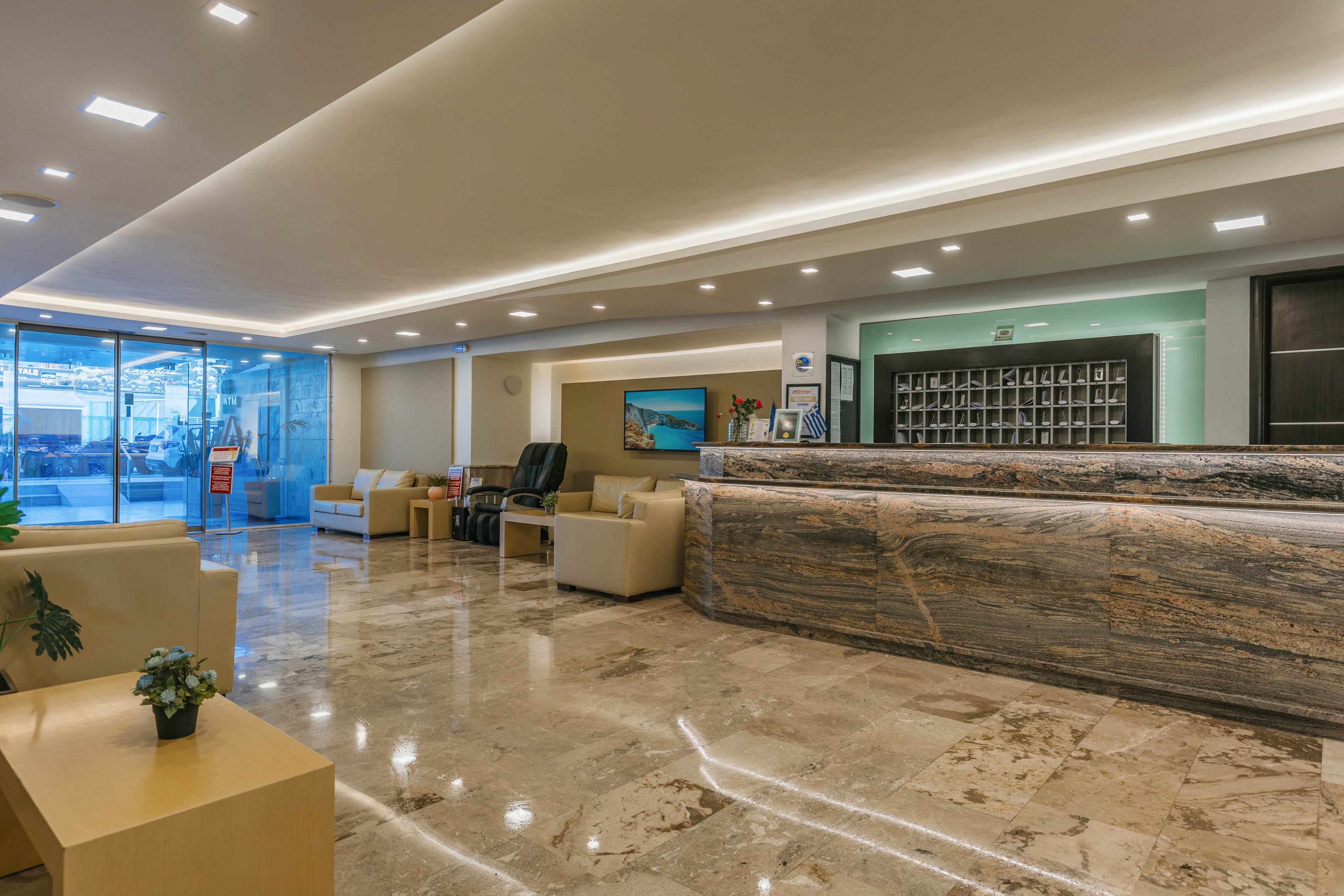 A spacious hotel lobby features a long marble reception desk as the central focus. Comfortable beige sofas and armchairs are arranged in seating areas on the polished stone floor. Large glass doors at the far end allow natural light to brighten the contemporary interior. Recessed ceiling lights and subtle decorative plants contribute to a clean, welcoming atmosphere.