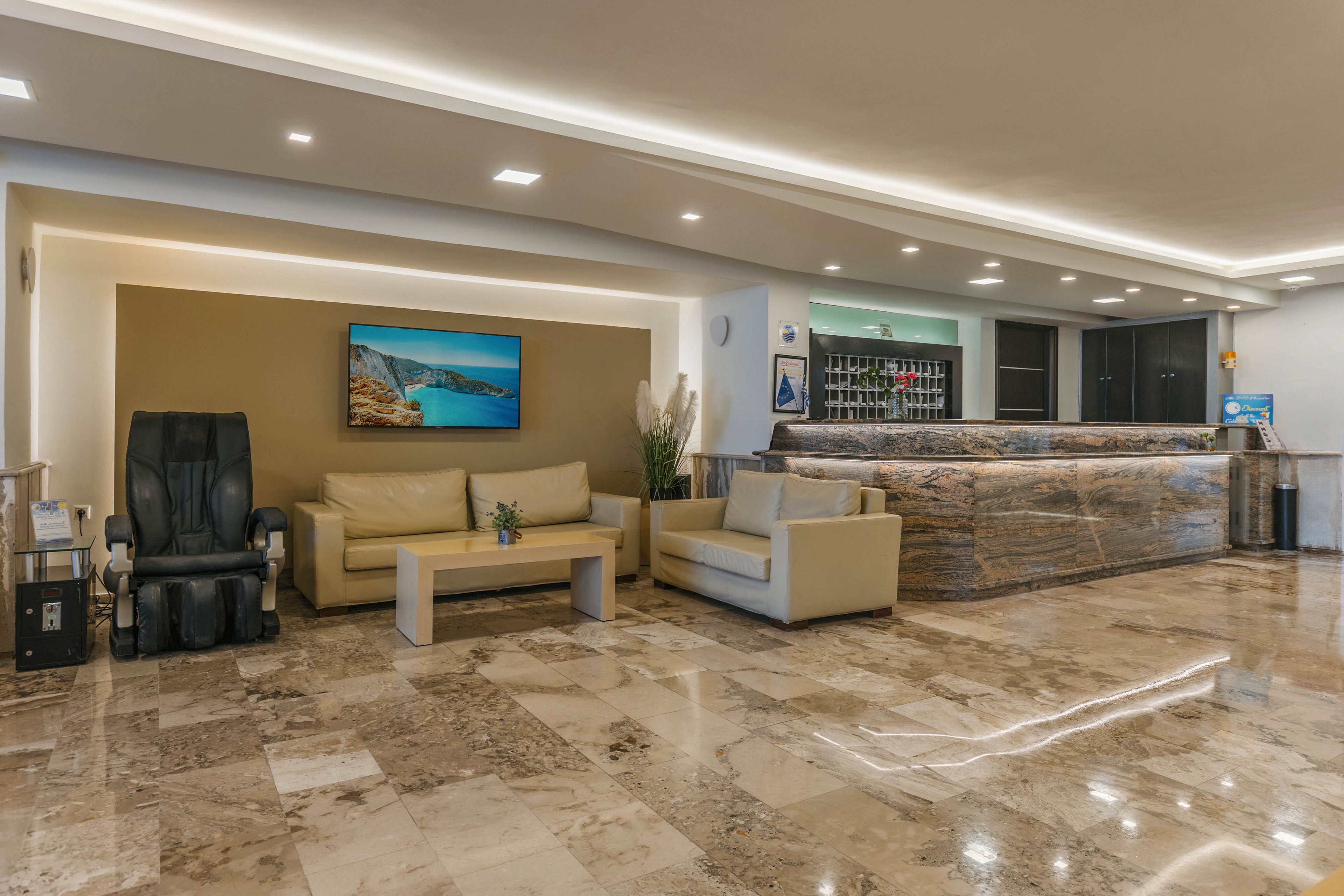 A contemporary hotel lobby features a polished marble floor and a long marble reception desk. Comfortable beige sofas and a matching coffee table create a seating area in front of a decorative wall. A large massage chair sits to the side, adding a touch of relaxation to the space. Warm recessed lighting and a landscape artwork on the wall contribute to a calm, welcoming atmosphere.
