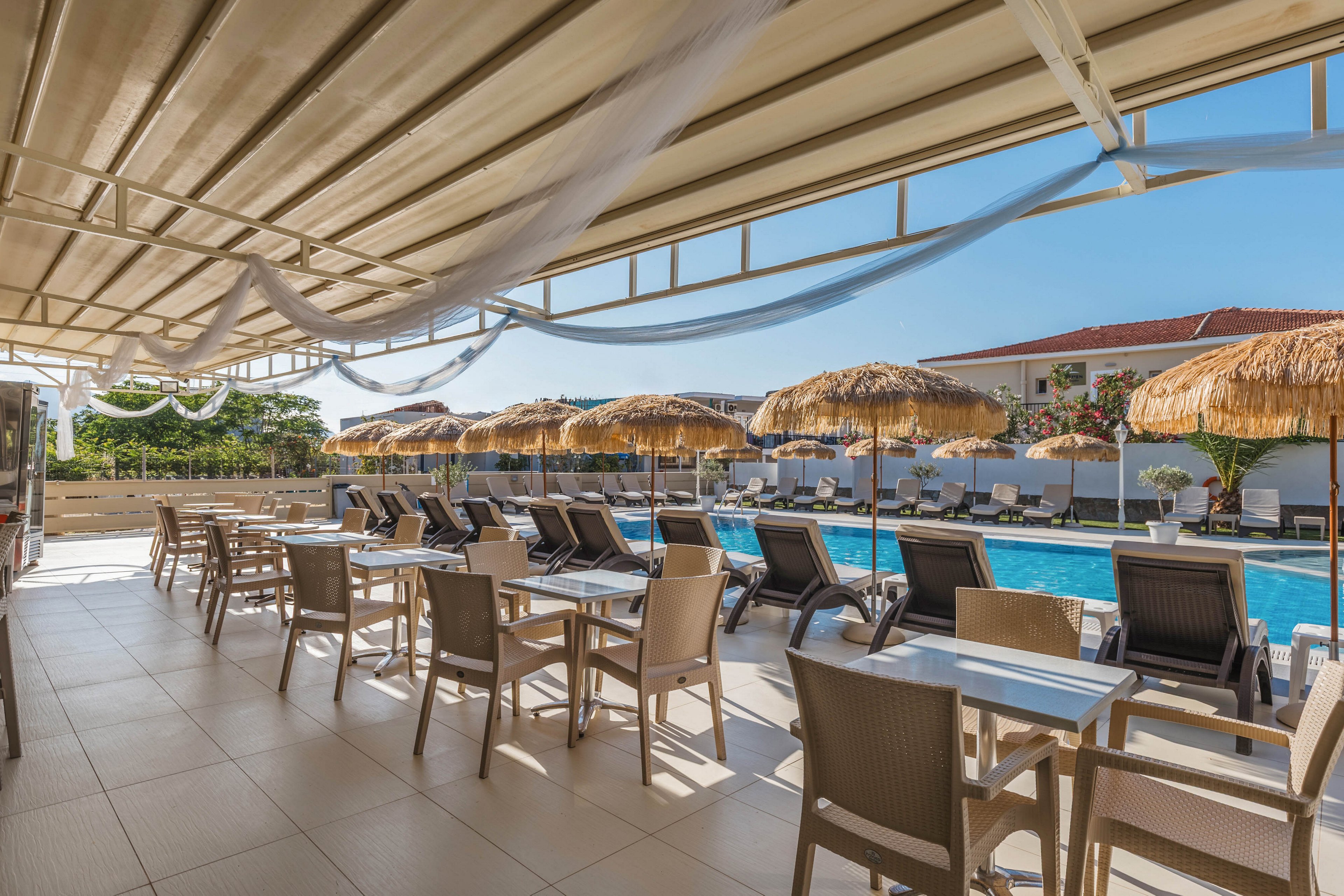 An outdoor resort terrace overlooks a swimming pool lined with sun loungers and straw parasols. Neatly arranged tables and chairs sit under a covered patio, providing shaded seating beside the water. The scene is bright and sunny, with clear blue sky and modern buildings in the background. The overall atmosphere suggests a relaxing vacation setting at a hotel or resort.
