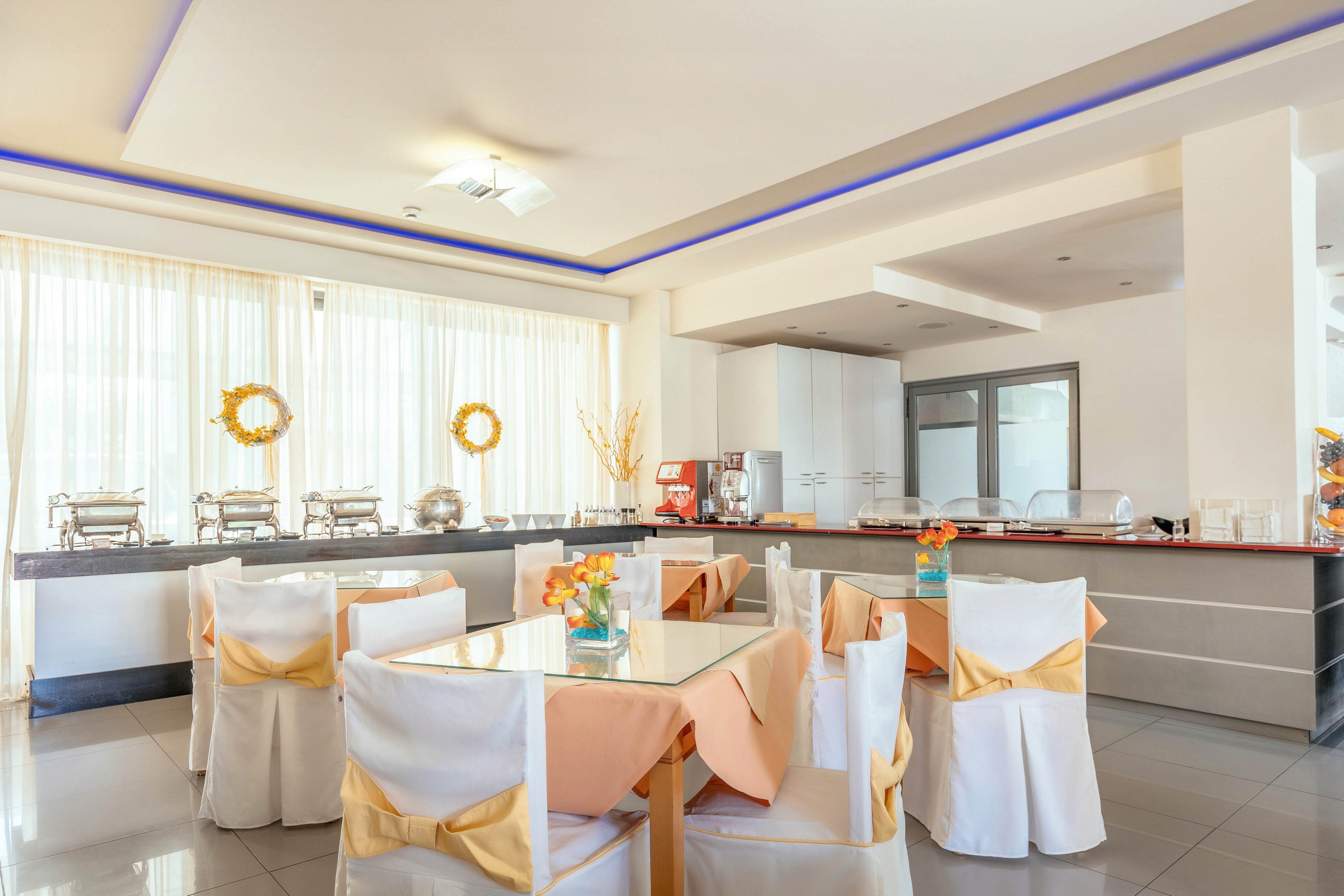 A bright banquet hall is set up with round tables covered in white cloths and peach table runners. Each chair is wrapped in white fabric and tied with a yellow sash, creating a coordinated event look. Along the back wall, a buffet counter holds covered chafing dishes and decorative elements. Large windows with sheer curtains allow natural light to fill the modern, clean interior.