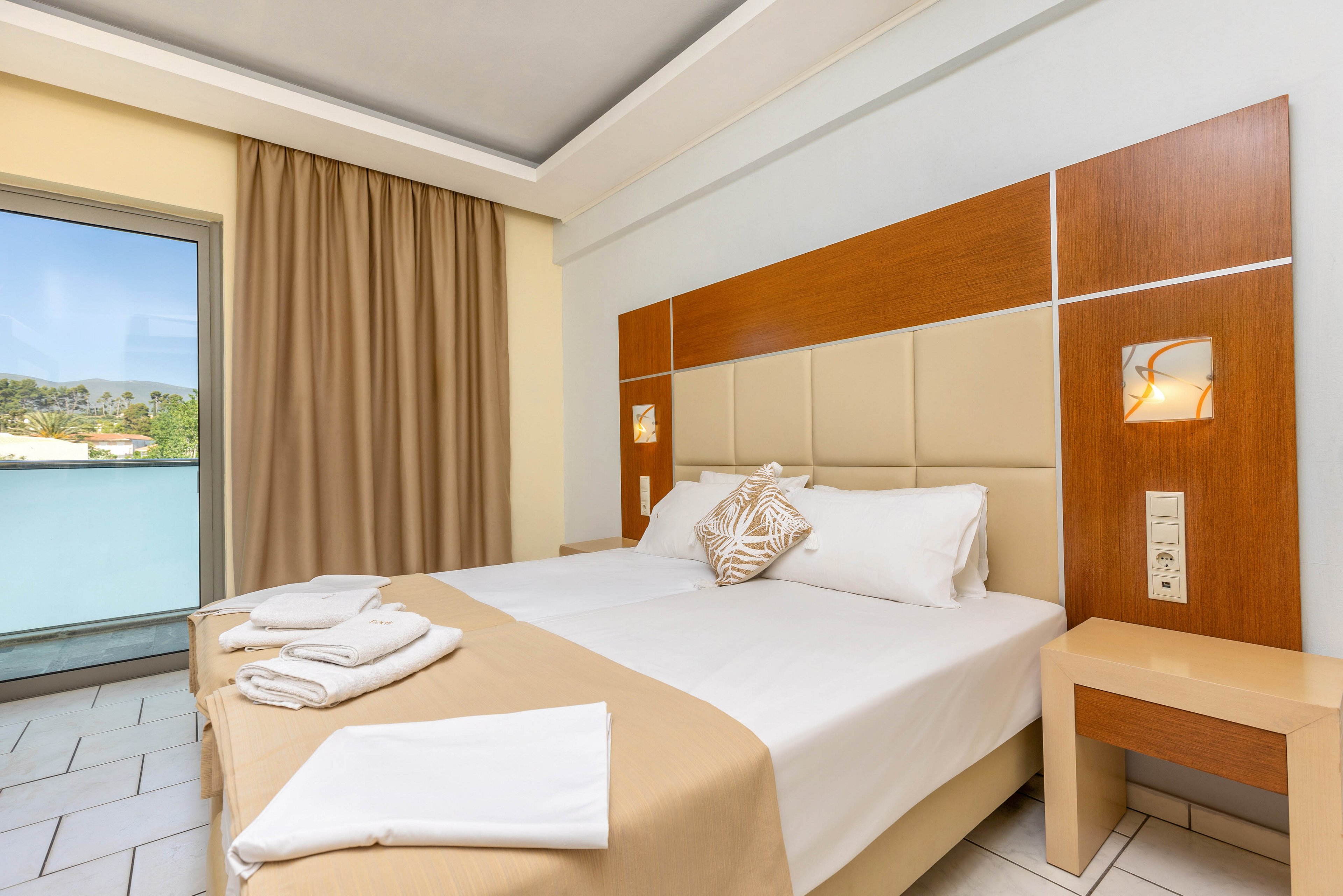A neatly made double bed with white linens and beige accents is the focal point of this modern hotel room. The space features a wooden and upholstered headboard with built-in reading lights and a small bedside table. Freshly folded towels are placed on the bed, suggesting readiness for guests. A large window with beige curtains opens to a balcony, allowing natural light to brighten the clean, minimalist interior.