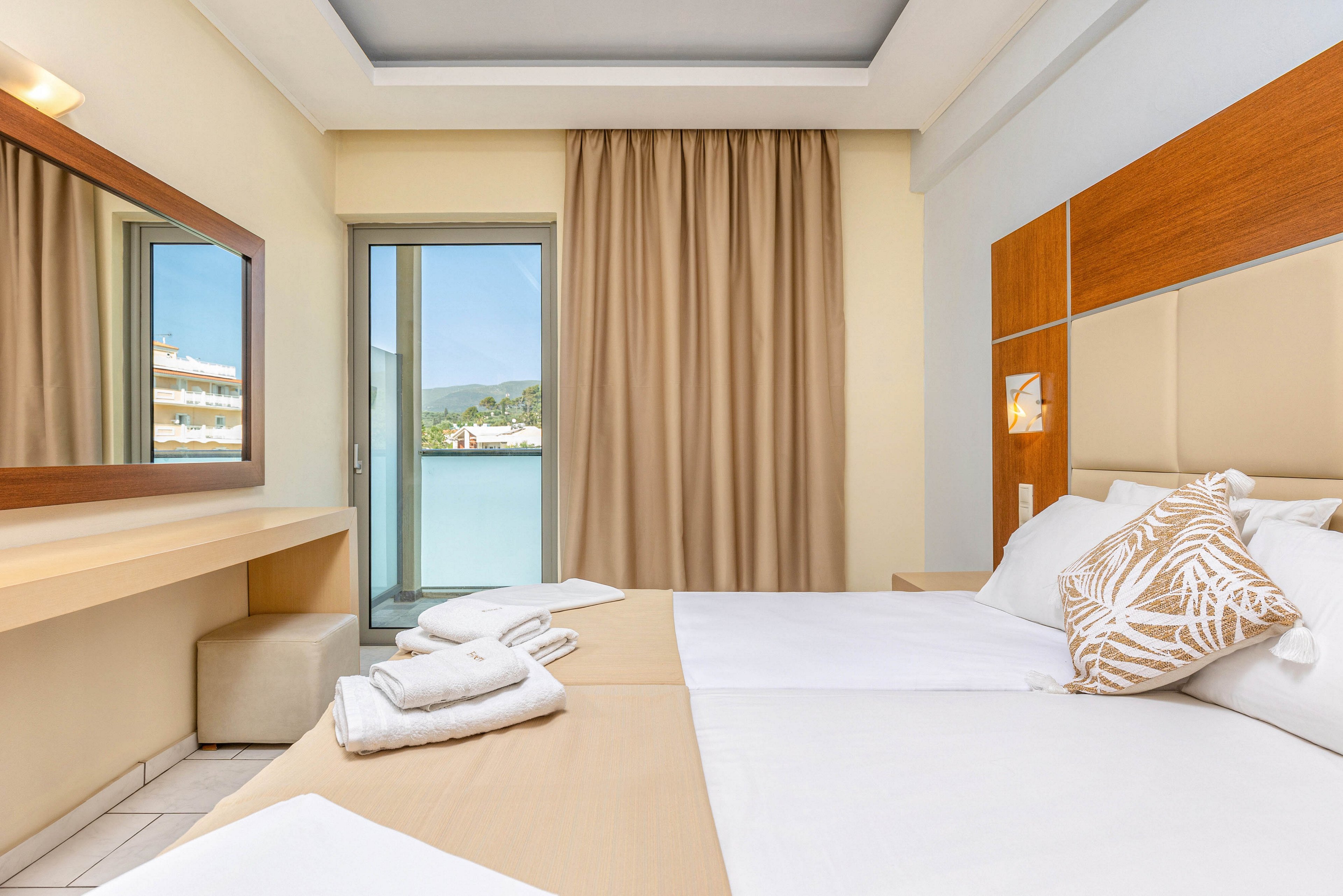 A neatly made hotel bedroom features two single beds pushed together with white linens and a decorative pillow. The room includes a light wood headboard, a wall mirror above a minimalist desk, and a small upholstered stool. Beige curtains frame a glass door that opens to a balcony with a view of water and distant buildings. Soft neutral tones and clean lines create a calm, contemporary atmosphere.