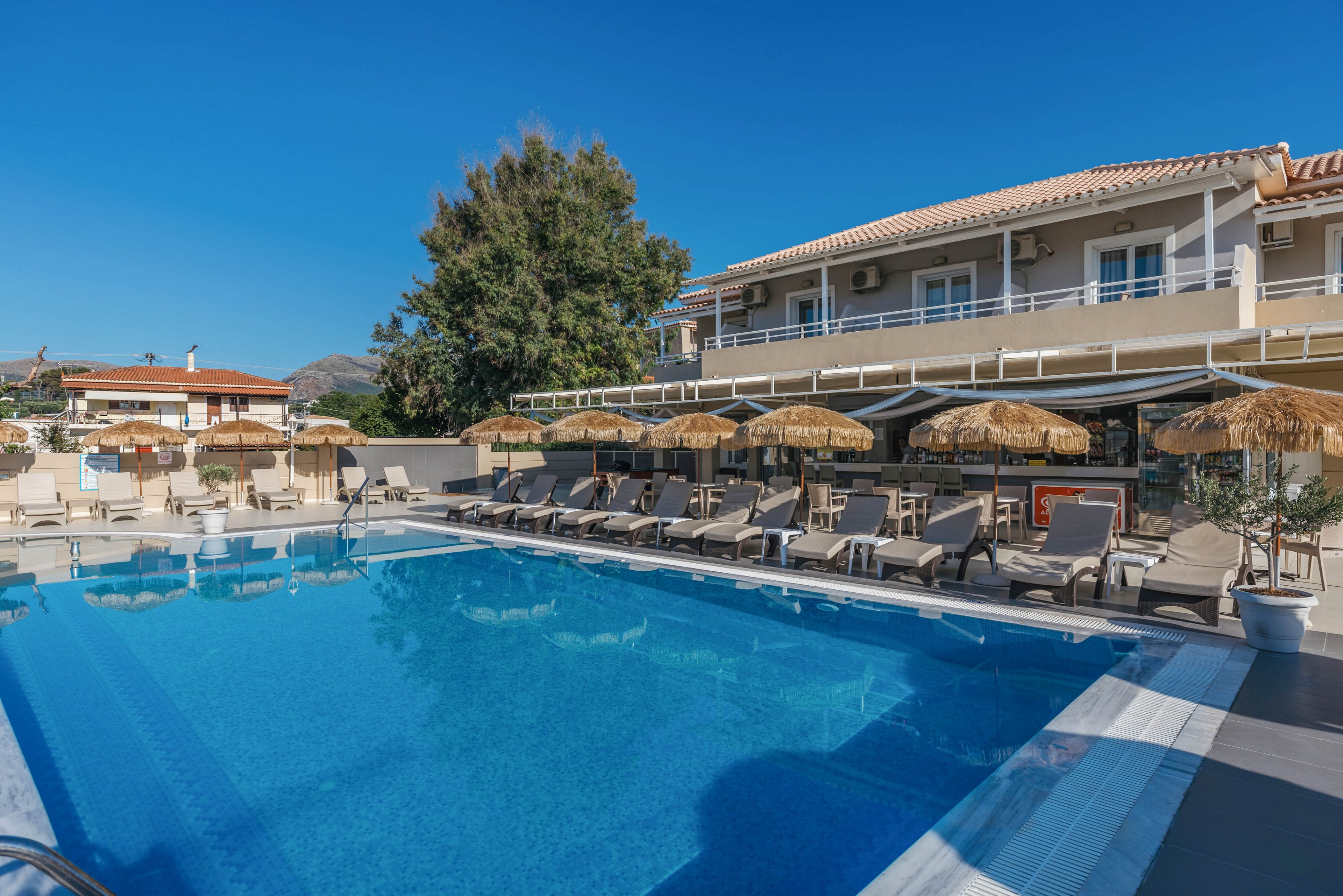 A clear blue swimming pool sits in front of a modern resort building under a bright sky. Numerous sun loungers and straw parasols line the poolside terrace, creating a relaxing holiday atmosphere. The two-story structure features balconies, large windows, and shaded outdoor seating areas. Nearby trees and distant hills frame the scene, emphasizing a tranquil vacation setting.