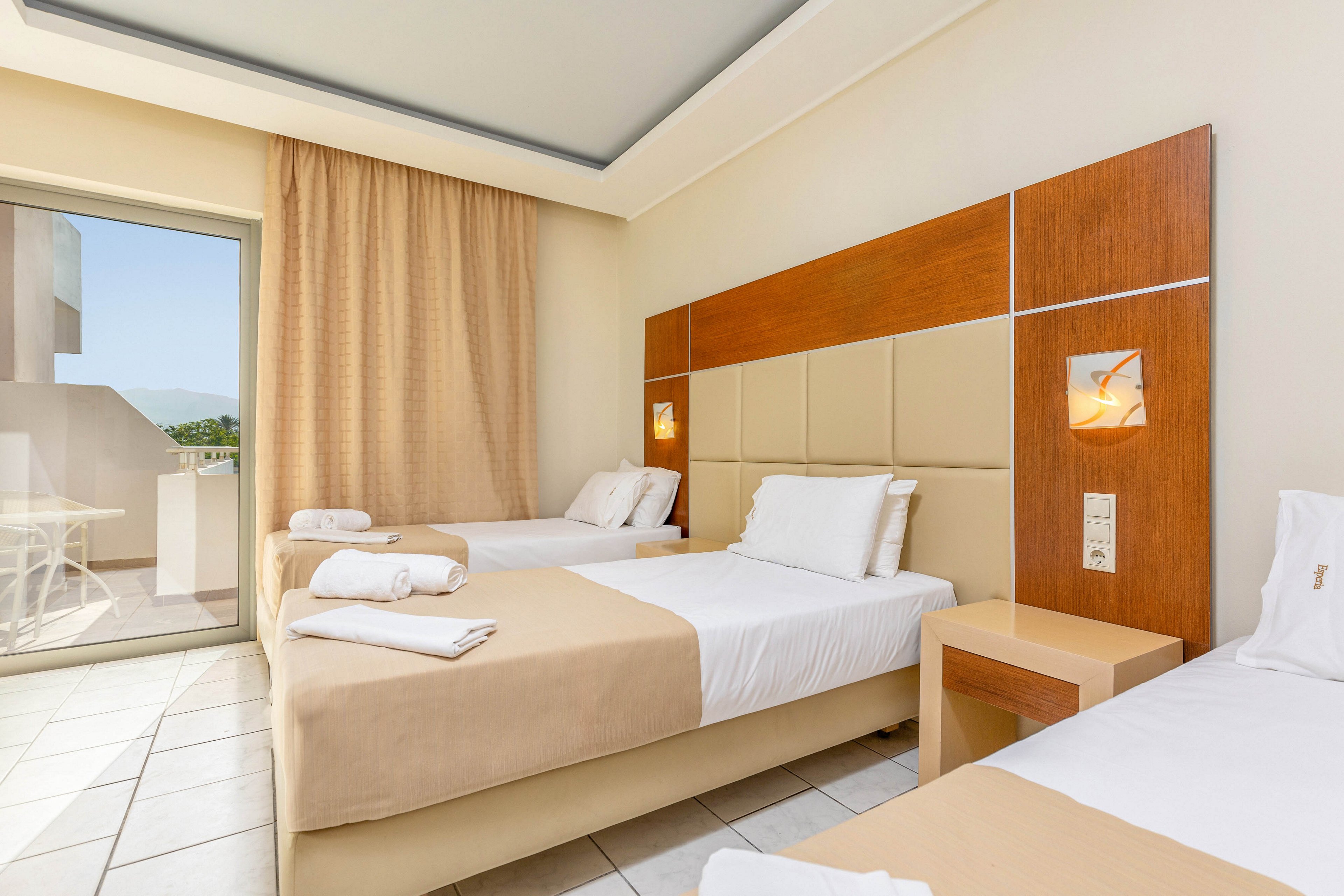 A bright, modern hotel room features two neatly made single beds with beige bed runners and white linens. A third bed is partially visible in the foreground, all aligned against a padded headboard with wooden accents. Warm wall lights and floor-to-ceiling curtains create a cozy atmosphere. A sliding glass door opens to a balcony with outdoor seating and a view of distant greenery.